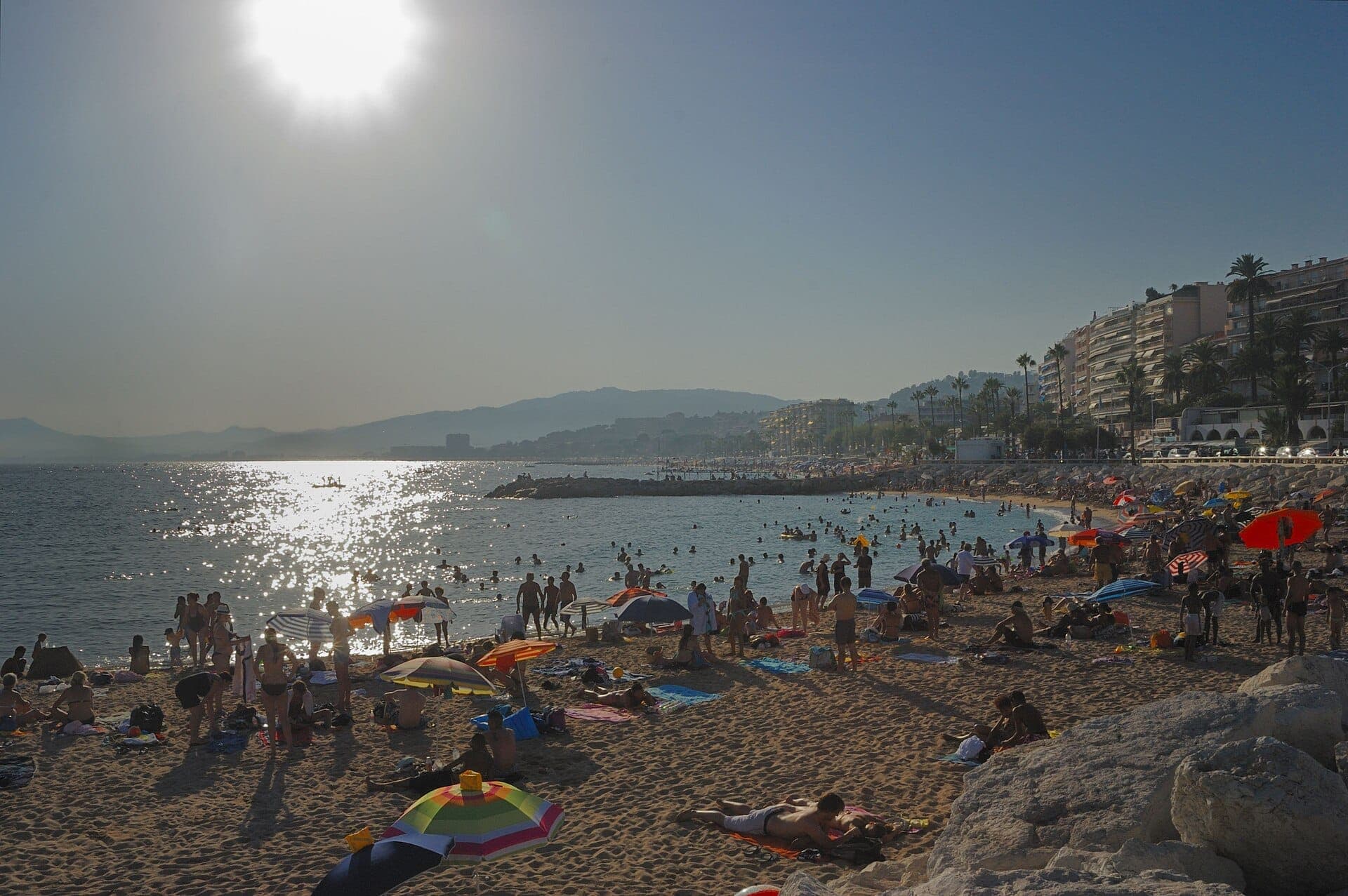 Многолюдный пляж Plage du Midi в Каннах с загорающими, разноцветными зонтиками, сверкающей водой и жилыми домами вдоль солнечного берега.