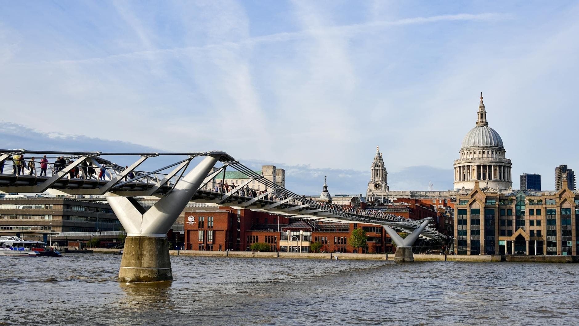 Cầu Millennium London bắc qua sông Thames với nhiều người đang qua lại, Nhà thờ St Paul's và các tòa nhà xung quanh hiện rõ phía xa dưới bầu trời xanh.