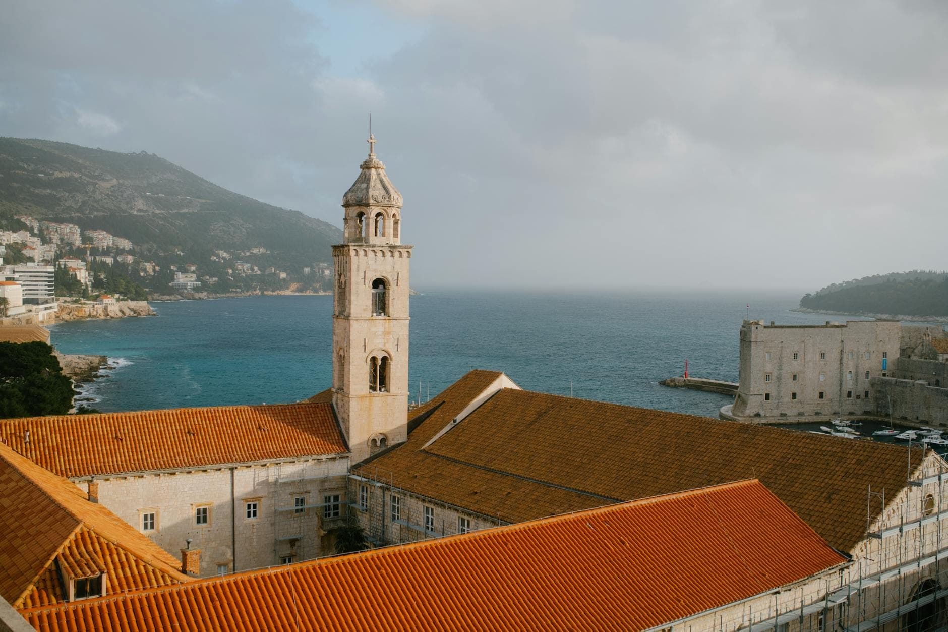 Blick von den Stadtmauern Dubrovniks auf den Minčeta-Turm über roten Ziegeldächern, mit dramatischer Meeres- und Küstenlandschaft im Hintergrund unter einem stimmungsvollen Himmel.
