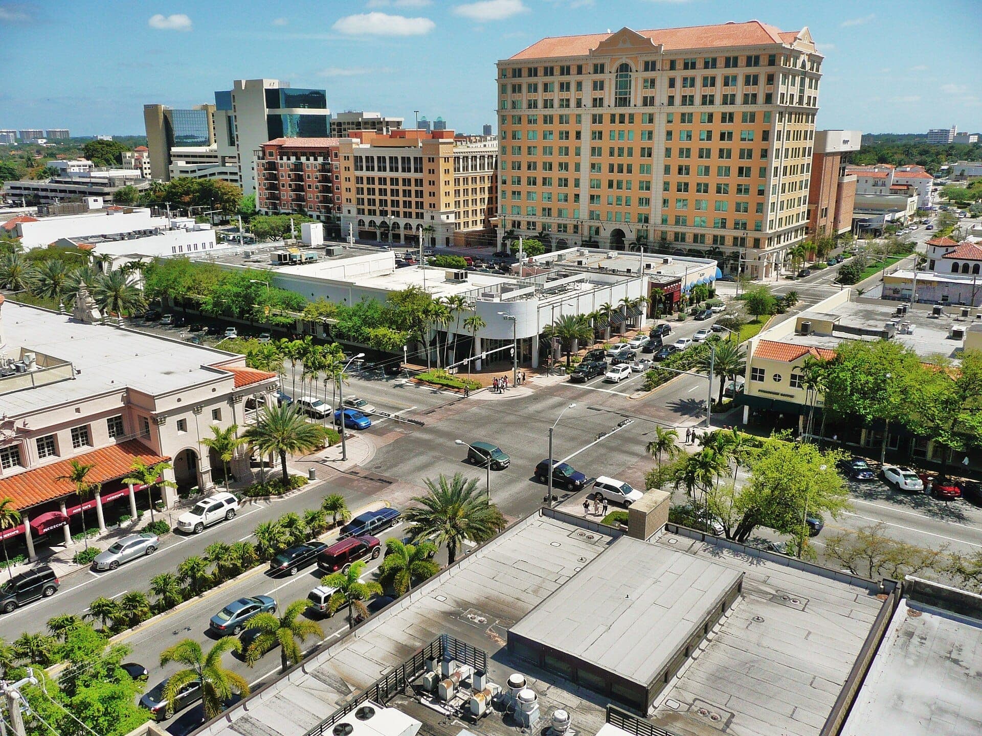 Vue aérienne de Miracle Mile à Coral Gables avec des palmiers, des bâtiments de style méditerranéen, des rues animées et une grande tour beige sous un ciel ensoleillé.