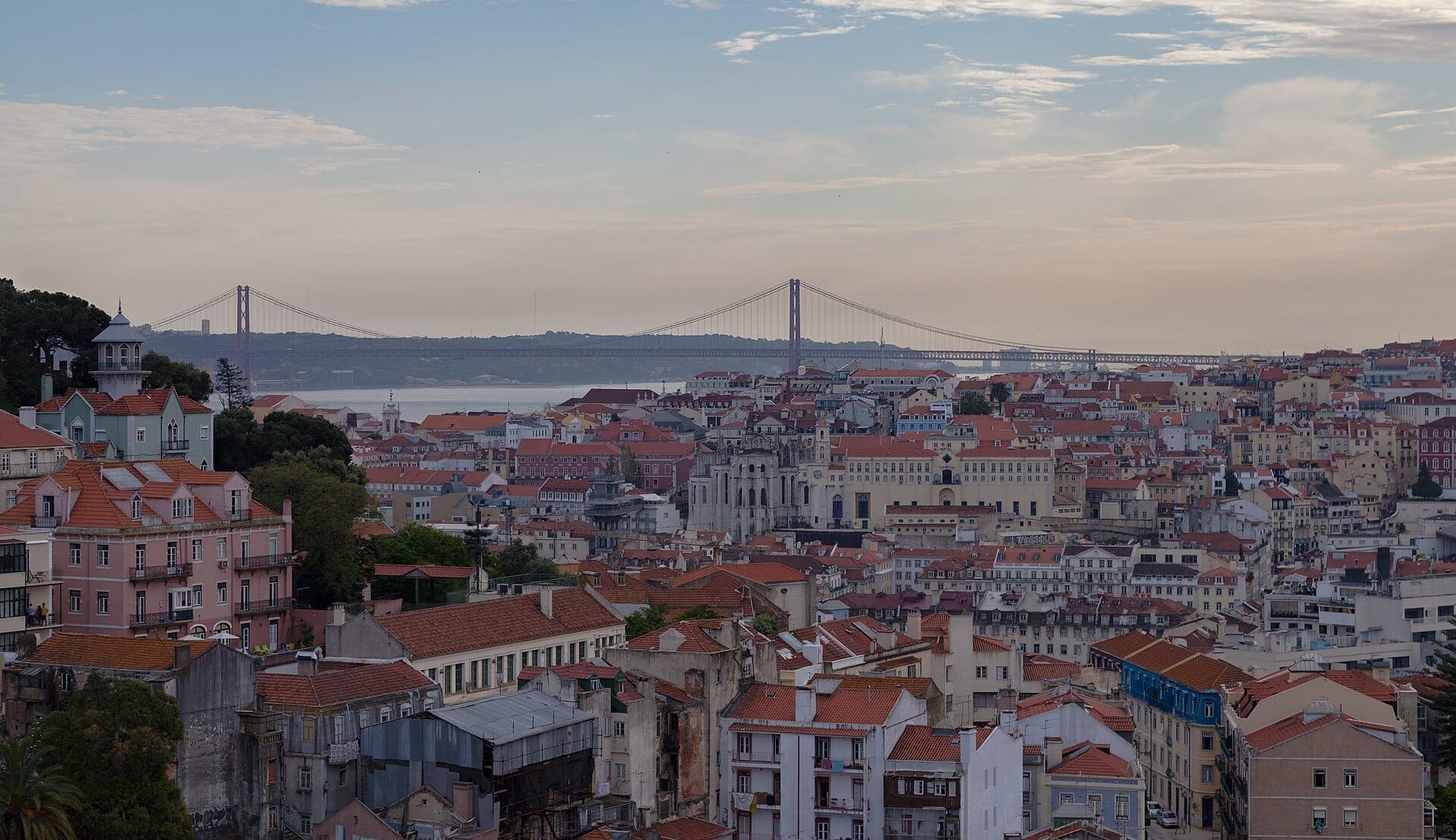 Miradouro da Graça com vista para os telhados vermelhos de Lisboa, edifícios históricos, o rio Tejo e a icónica Ponte 25 de Abril ao entardecer.
