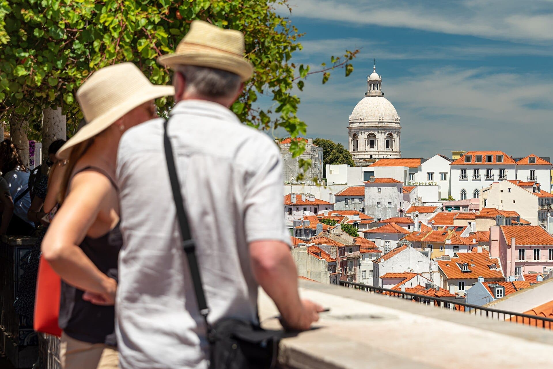 Visitantes no Miradouro de Santa Luzia contemplam os telhados alaranjados da Alfama e a cúpula branca do Panteão Nacional num dia ensolarado.