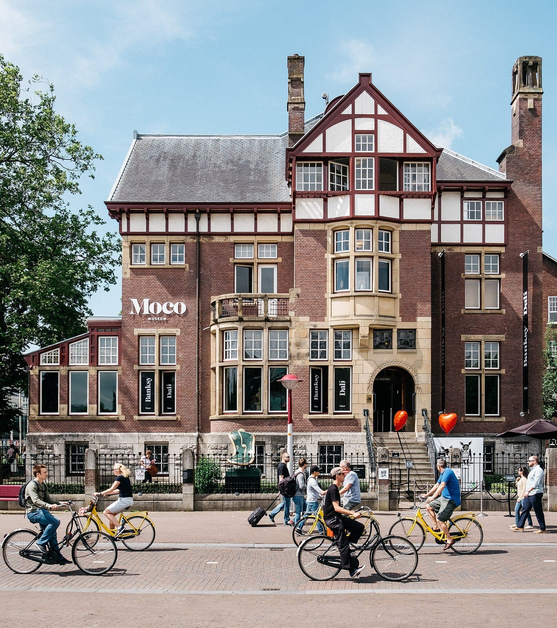 Das Moco Museum Amsterdam, eine historische Backsteinvilla mit großen Fenstern, steht an einem sonnigen Tag hinter Radfahrern und Fußgängern in der Nähe des Museumplein.