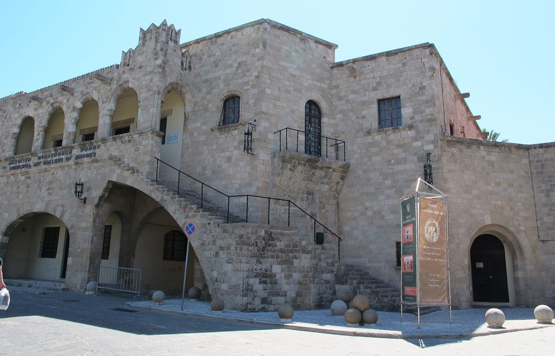Façade en pierre du Museum of Modern Greek Art à Rhodes, avec des fenêtres en arc, un escalier extérieur et un panneau du musée en plein jour.