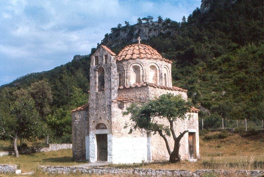 Extérieur en pierre du Monastère de Fountoukli, avec une architecture byzantine et un dôme à tuiles rouges, entouré de collines verdoyantes.