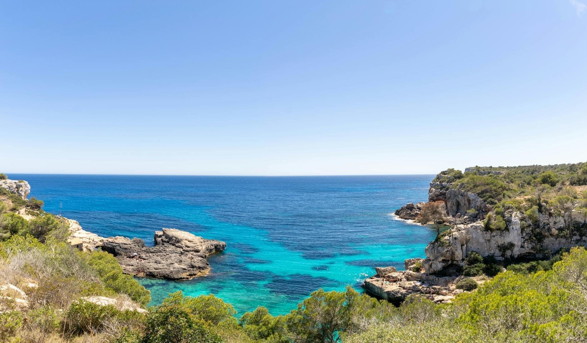 Kristallklares türkisfarbenes Wasser, gerahmt von Felsenklippen und üppig grünem Gebüsch unter strahlend blauem Himmel – die geschützte Küste des Naturparks Mondragó.