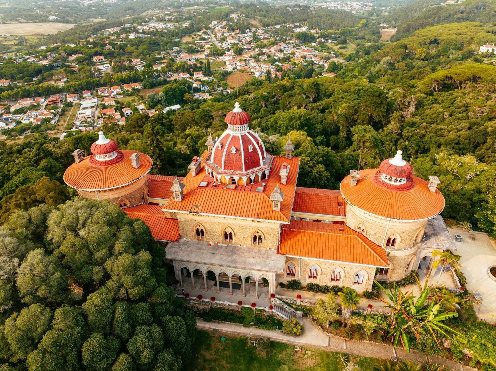 Vista aérea do Palácio de Monserrate cercado por jardins exuberantes, com suas cúpulas características e arquitetura rica em detalhes se destacando entre o verde de Sintra.
