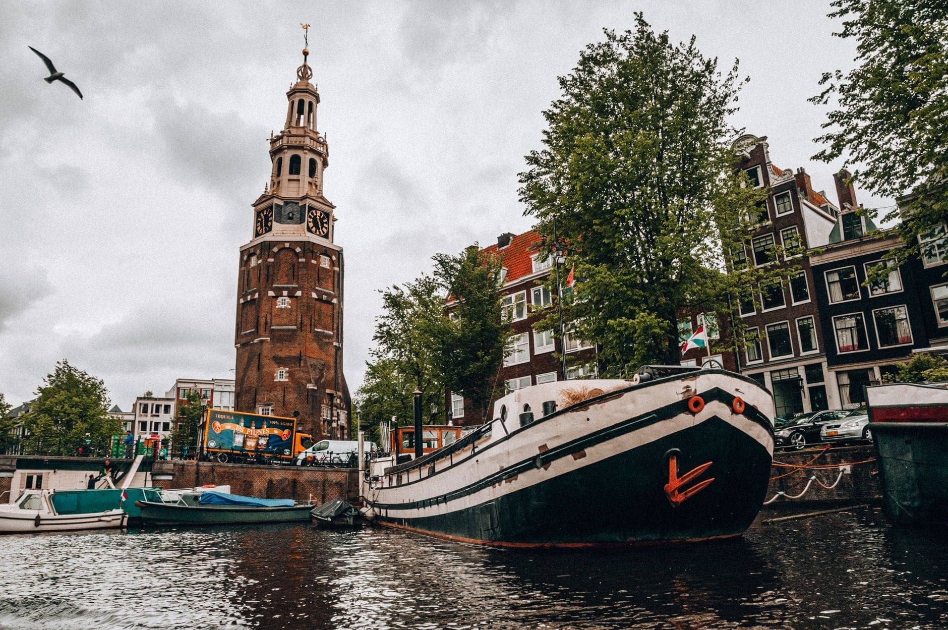 Blick auf den Montelbaanstoren am Oudeschans-Kanal in Amsterdam mit Hausbooten, Bäumen, Kanalhäusern und einem Vogel in einem bewölkten Himmel.
