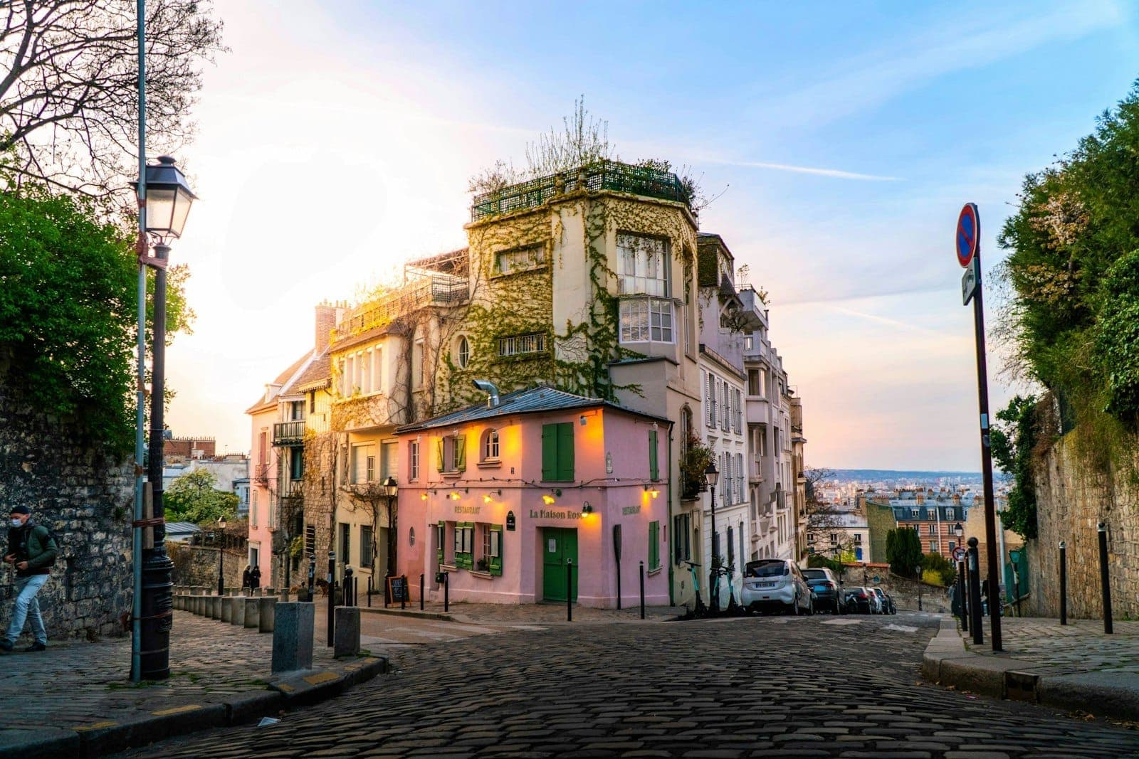 Charmante ruelle pavée de Montmartre avec des immeubles couverts de vigne, des façades pastel et un café rose baigné dans la douce lumière du soir, captant l'atmosphère bohème parisienne.