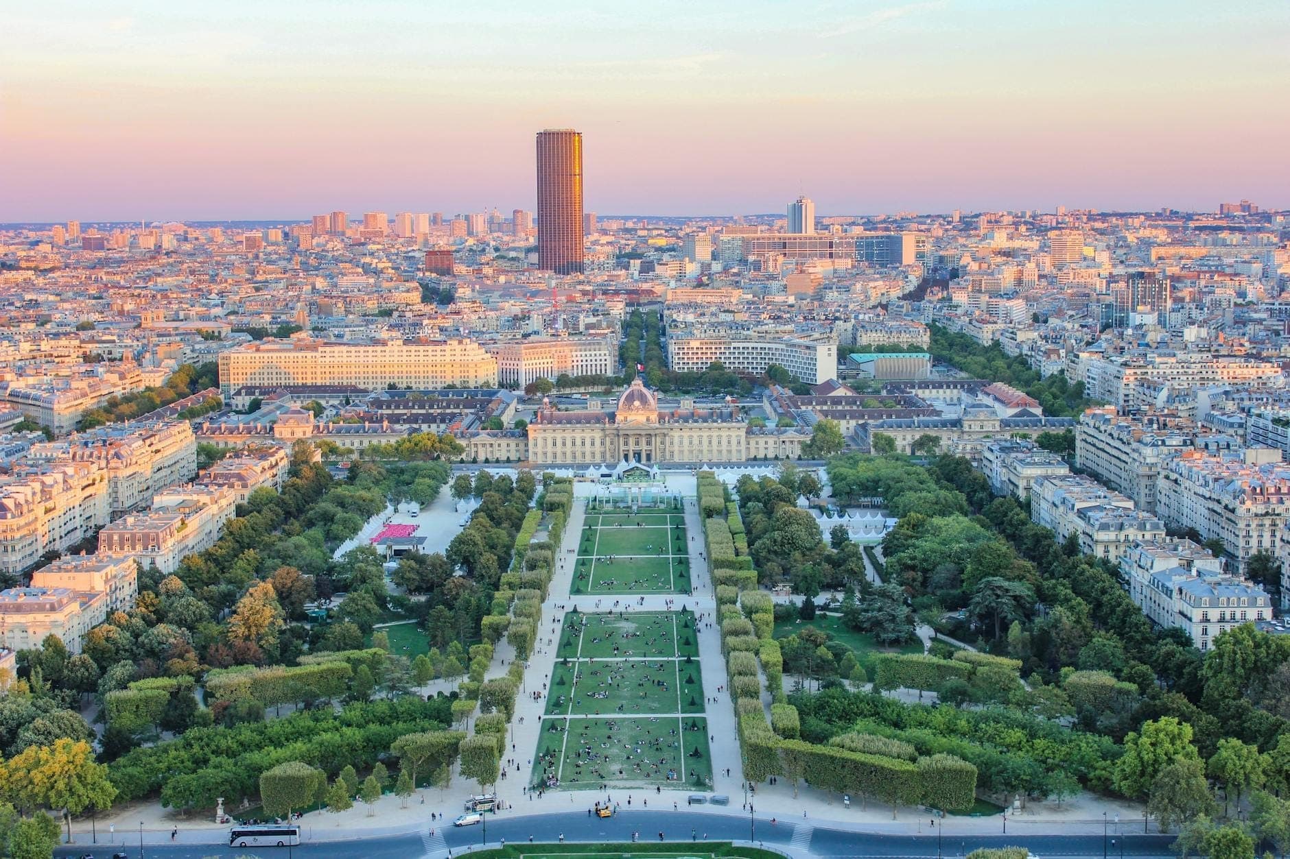 A panoramic view of Paris with the Tour Montparnasse tower centered in the background and the Champ de Mars park stretching out below at sunset.
