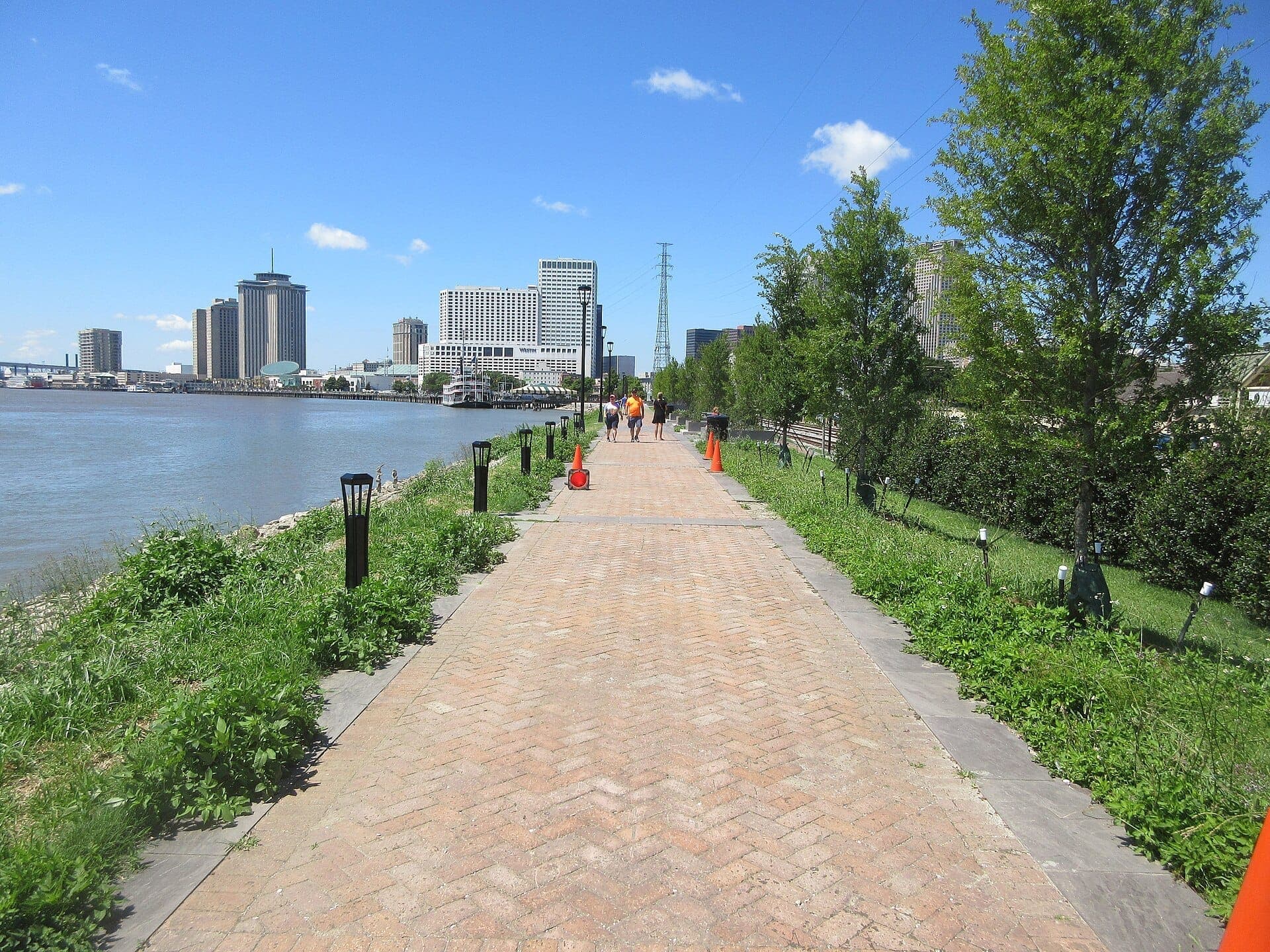 Brick-paved Moon Walk promenade with people walking, green trees, and views of downtown New Orleans skyline along the Mississippi River.