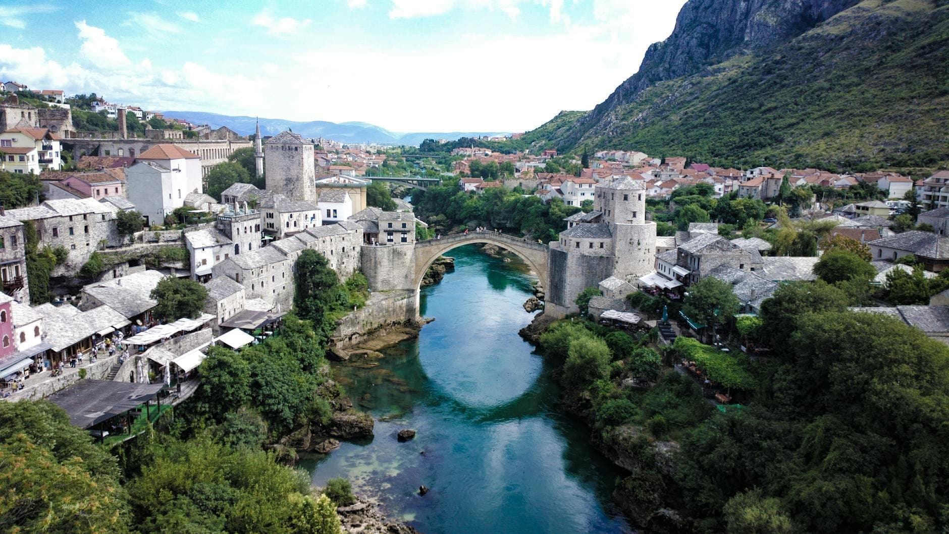 Weitläufige Luftaufnahme von Stari Most (Alte Brücke) über dem smaragdgrünen Fluss Neretva in Mostar, umgeben von historischen Steingebäuden und dramatischen grünen Bergen.