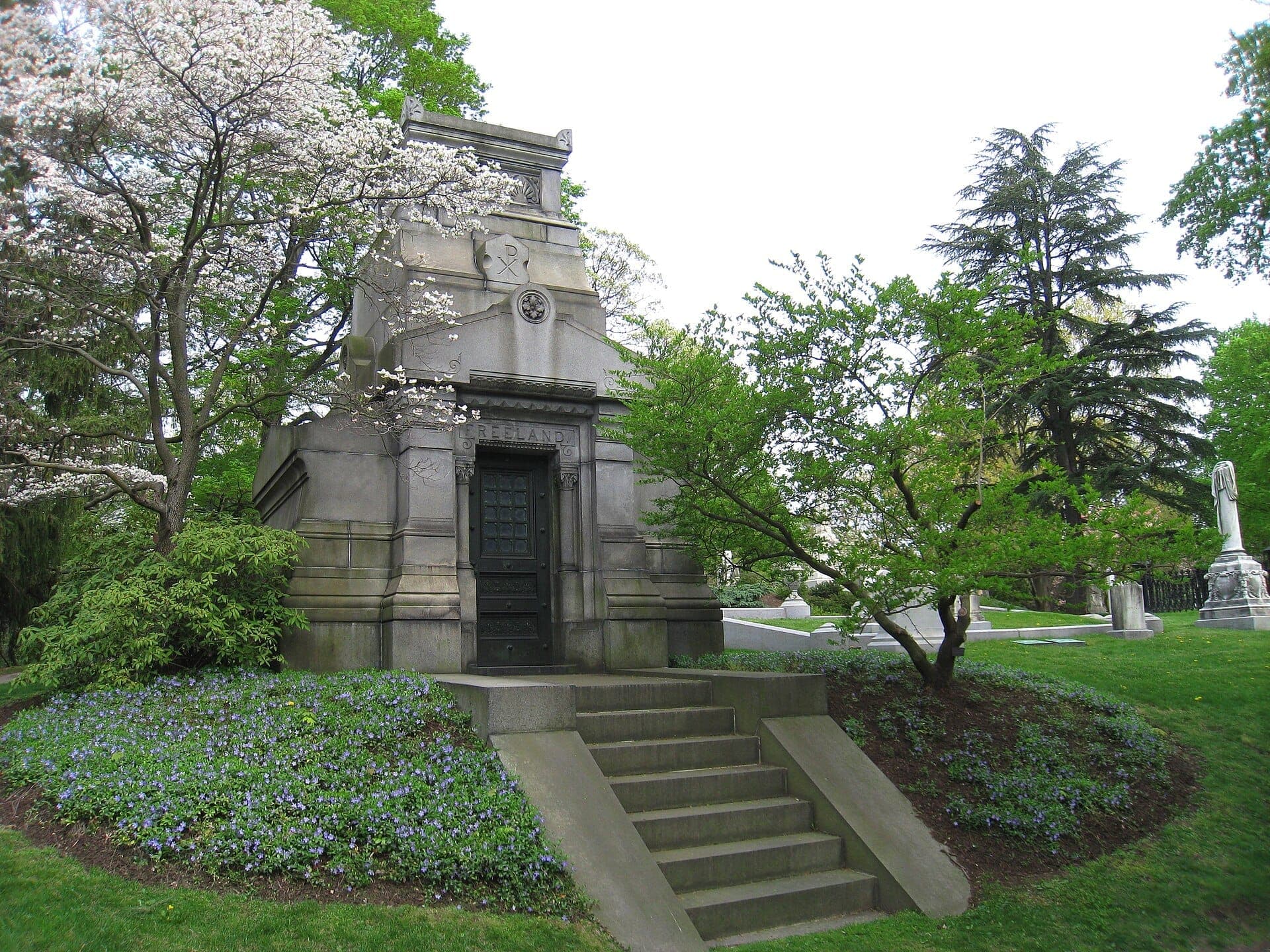Mausolée en pierre entouré de fleurs printanières, d'arbres luxuriants et de pierres tombales au cimetière de Mount Auburn par une journée lumineuse et dégagée.