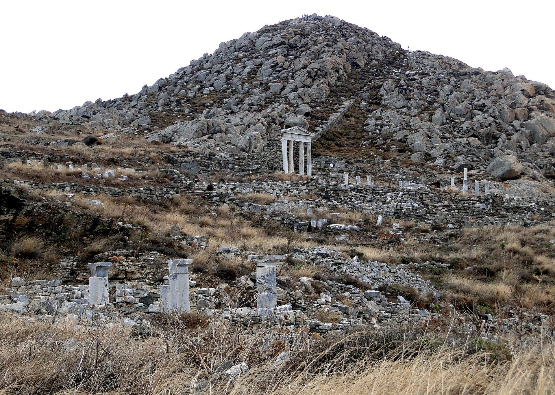 Felsige Hänge des Berges Kynthos auf der Insel Delos mit antiken Steinruinen und einem griechischen Tempel unter einem bedeckten Himmel.