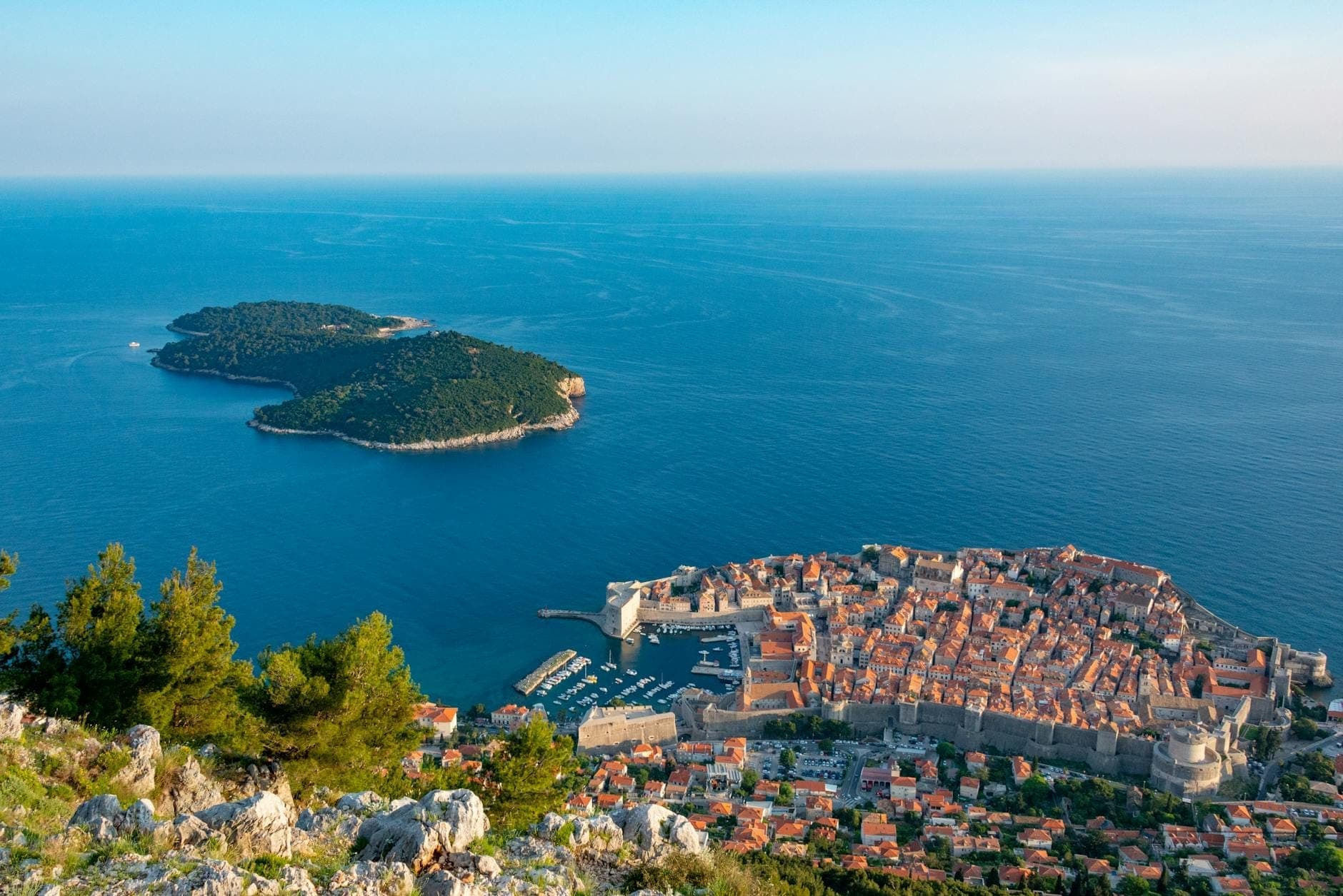 Vue imprenable depuis le mont Srđ sur la vieille ville de Dubrovnik, la mer Adriatique et l'île de Lokrum par une belle journée ensoleillée, avec des pins verts au premier plan.