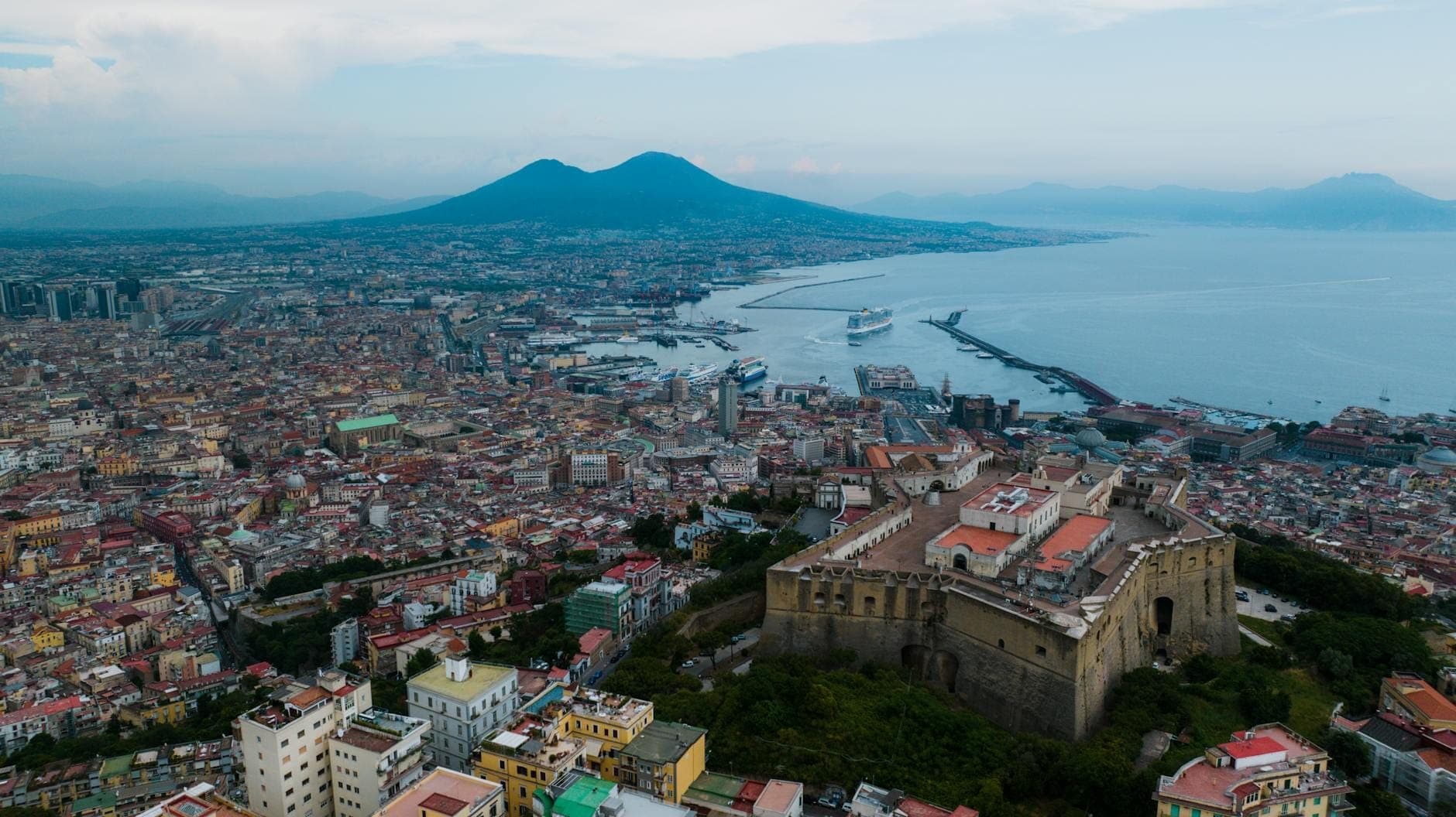 Uma vista aérea impressionante de Nápoles com o Monte Vesúvio ao fundo, a cidade movimentada e o porto em primeiro plano sob um céu vibrante.