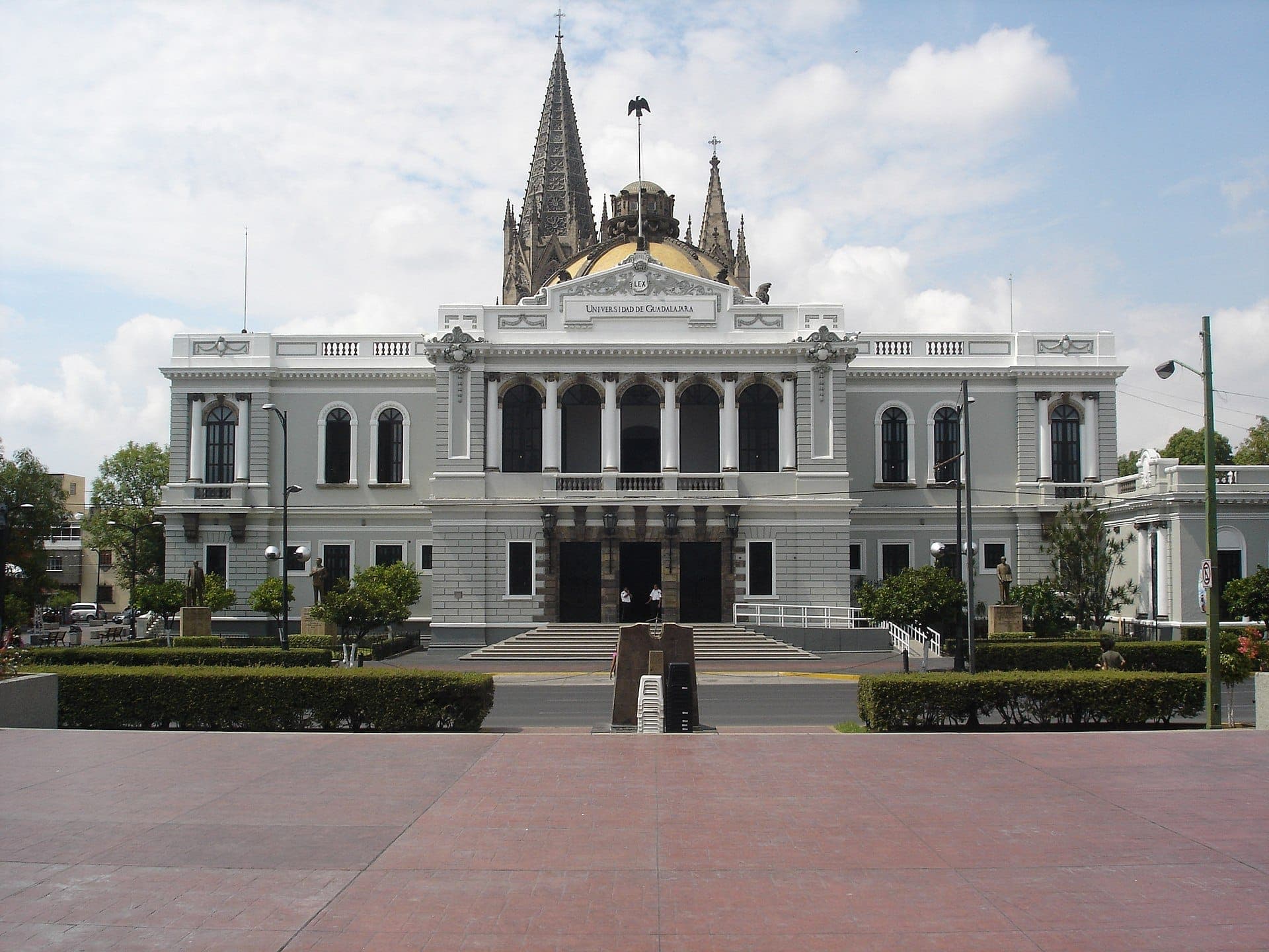 ด้านหน้าอาคารสไตล์นีโอคลาสสิกสีขาวประดับประดาของ MUSA, Museo de las Artes de la Universidad de Guadalajara พร้อมยอดแหลมและโดมภายใต้ท้องฟ้ามีเมฆบางส่วน
