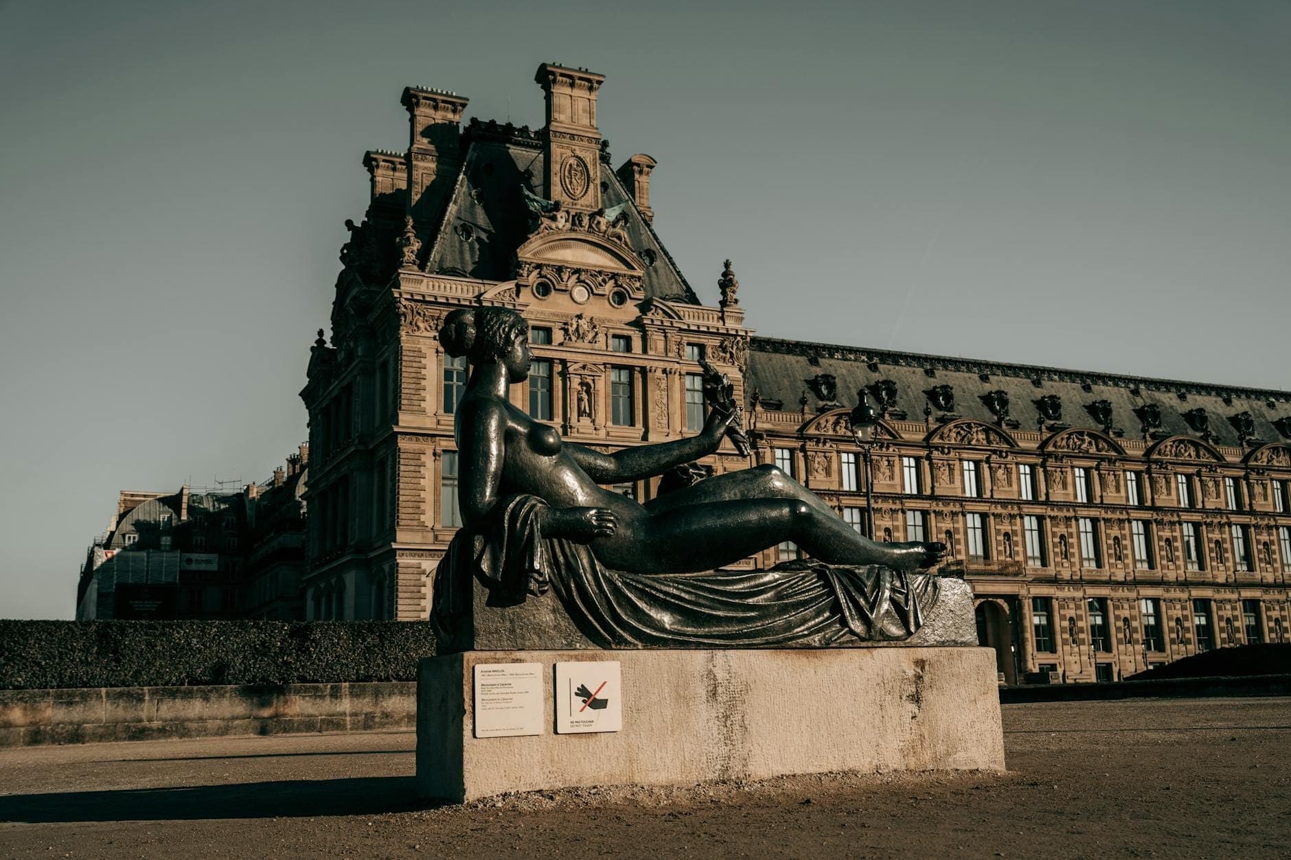 Large bronze reclining female sculpture set outdoors in front of ornate historic Parisian building under clear sky, evoking an artistic, open-air museum experience.