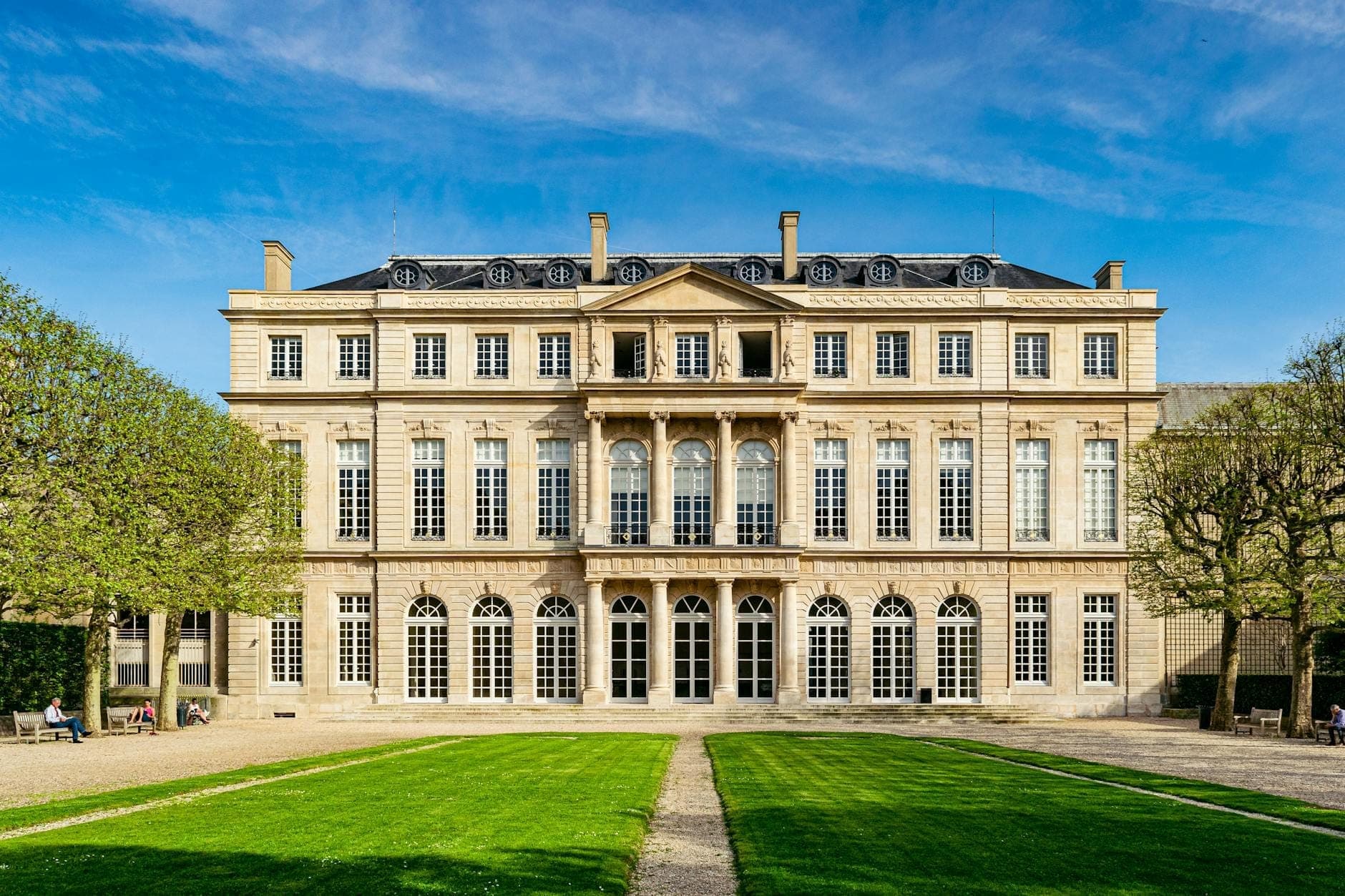 The elegant 16th-century facade of Musée Carnavalet with arched windows, manicured lawn, and blue sky, inviting visitors to explore Paris history.