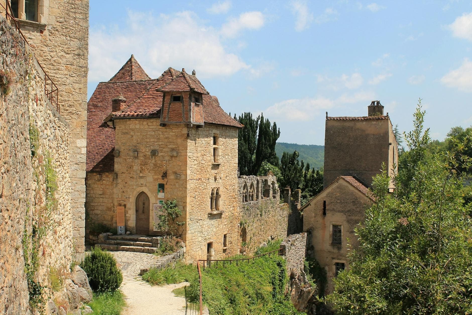 Picturesque medieval stone buildings with steep roofs and a narrow pathway, nestled on a sunny hillside with lush greenery.