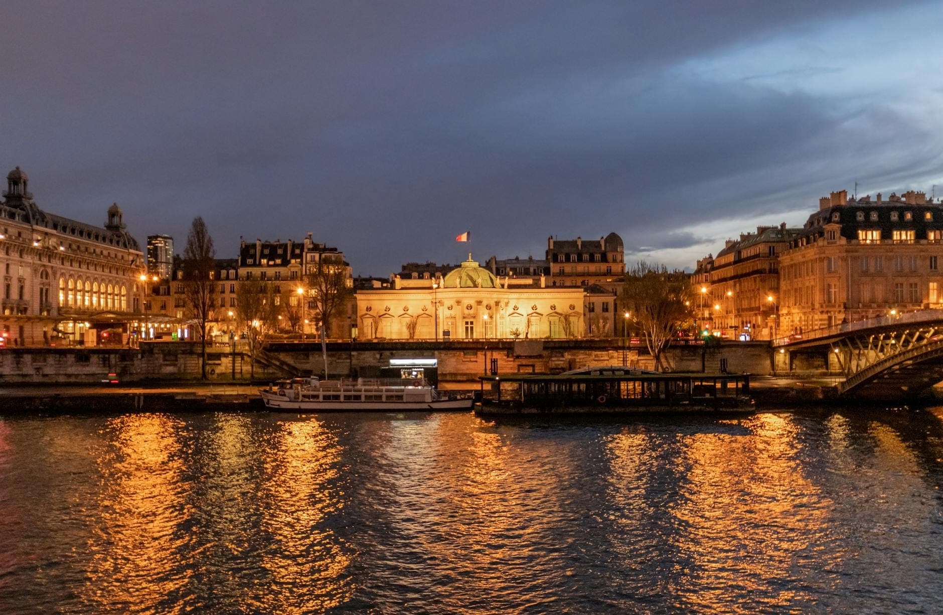 Elegant museum building lit up at dusk viewed from across the Seine, with glowing reflections on the river and Paris cityscape.