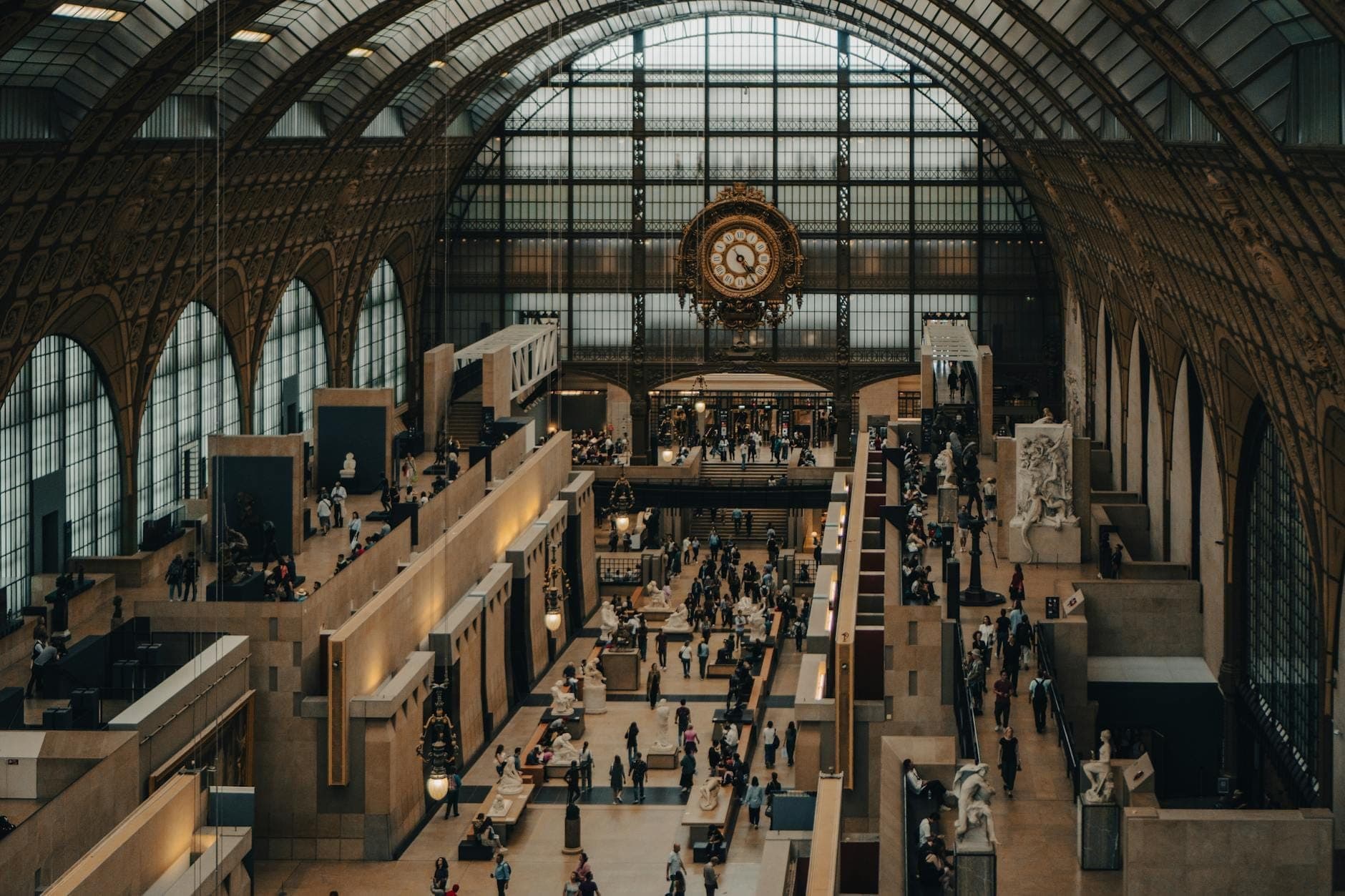 Wide interior view of Musée d'Orsay with its grand arched glass roof, iconic central clock, bustling visitors, and art sculptures on display.