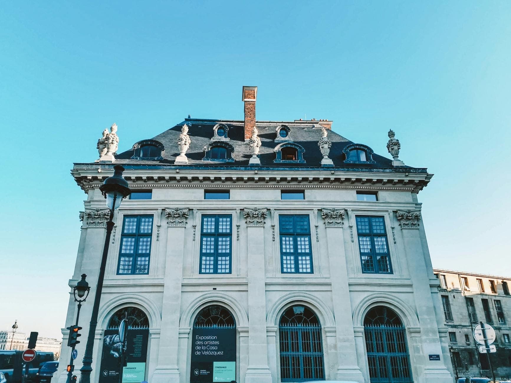Front facade of an elegant Parisian mansion with arched windows, ornate statues on the rooftop, and clear blue sky above.