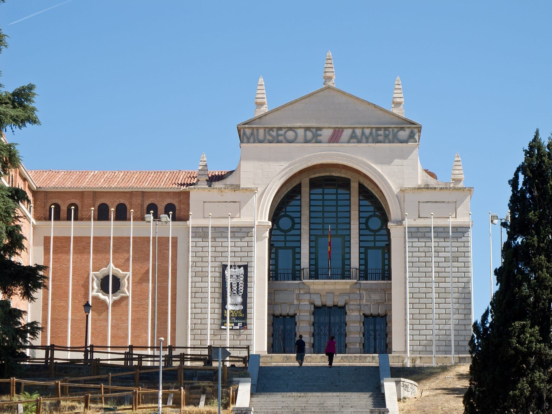 ทางเข้าหลักของ Museo de América ในมาดริด โดดเด่นด้วยด้านหน้าอาคารหินขนาดใหญ่ หน้าต่างโค้ง และผู้คนสองคนกำลังเดินขึ้นบันได