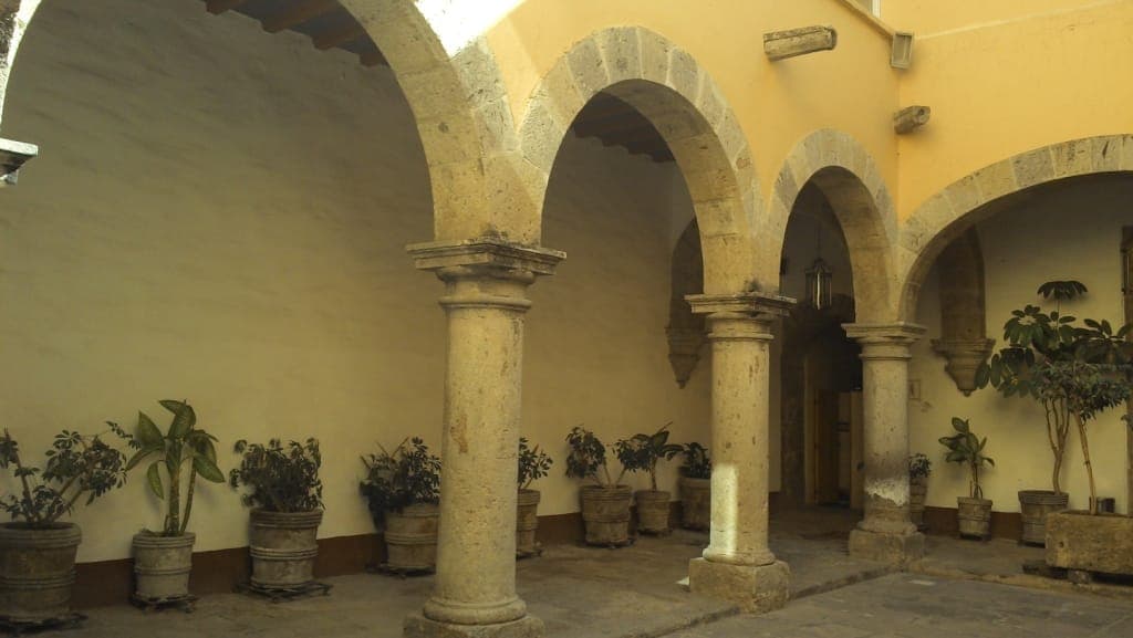 ซุ้มโค้งหินและต้นไม้กระถางเรียงรายในลานกว้างอันสงบของ Museo de la Ciudad de Guadalajara ภายใต้แสงยามบ่ายที่อบอุ่น