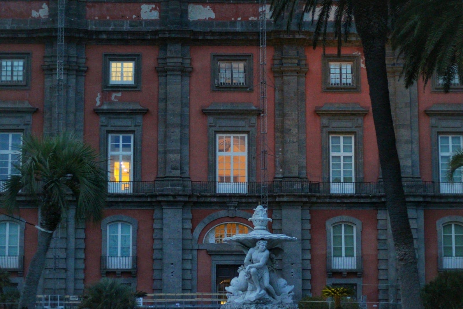 Vista frontal do Museo e Real Bosco di Capodimonte mostrando sua fachada vermelha, grandes janelas iluminadas por dentro e uma fonte clássica.