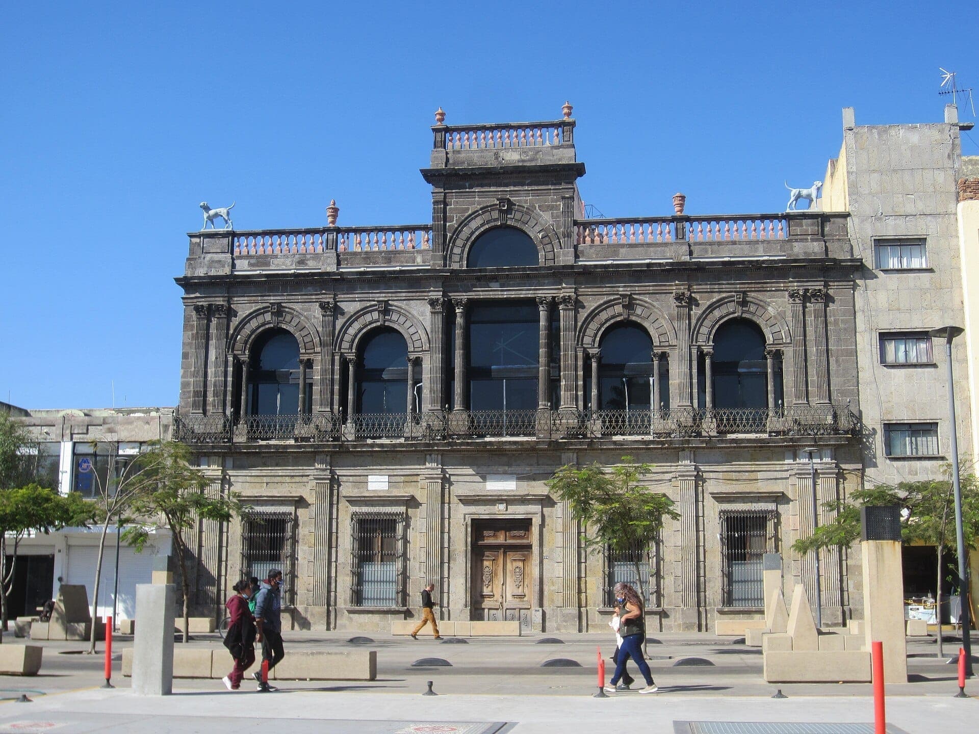 ด้านหน้าอาคารประวัติศาสตร์ของ Museo del Periodismo y las Artes Gráficas ในเมือง Guadalajara มีหน้าต่างโค้งอาร์ช งานหินแกะสลักประดับประดา และรูปปั้นสุนัขสองตัวบนหลังคา