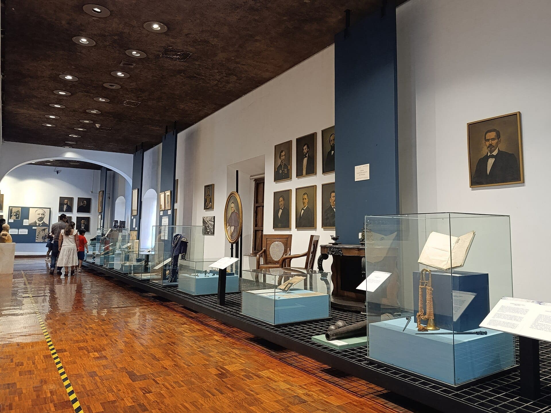 ห้องจัดแสดงนิทรรศการภายใน Museo Regional de Guadalajara พร้อมตู้กระจกโชว์ โบราณวัตถุทางประวัติศาสตร์ พื้นไม้ และภาพพอร์ตเทรตเรียงรายตามผนังสีน้ำเงินและสีขาว