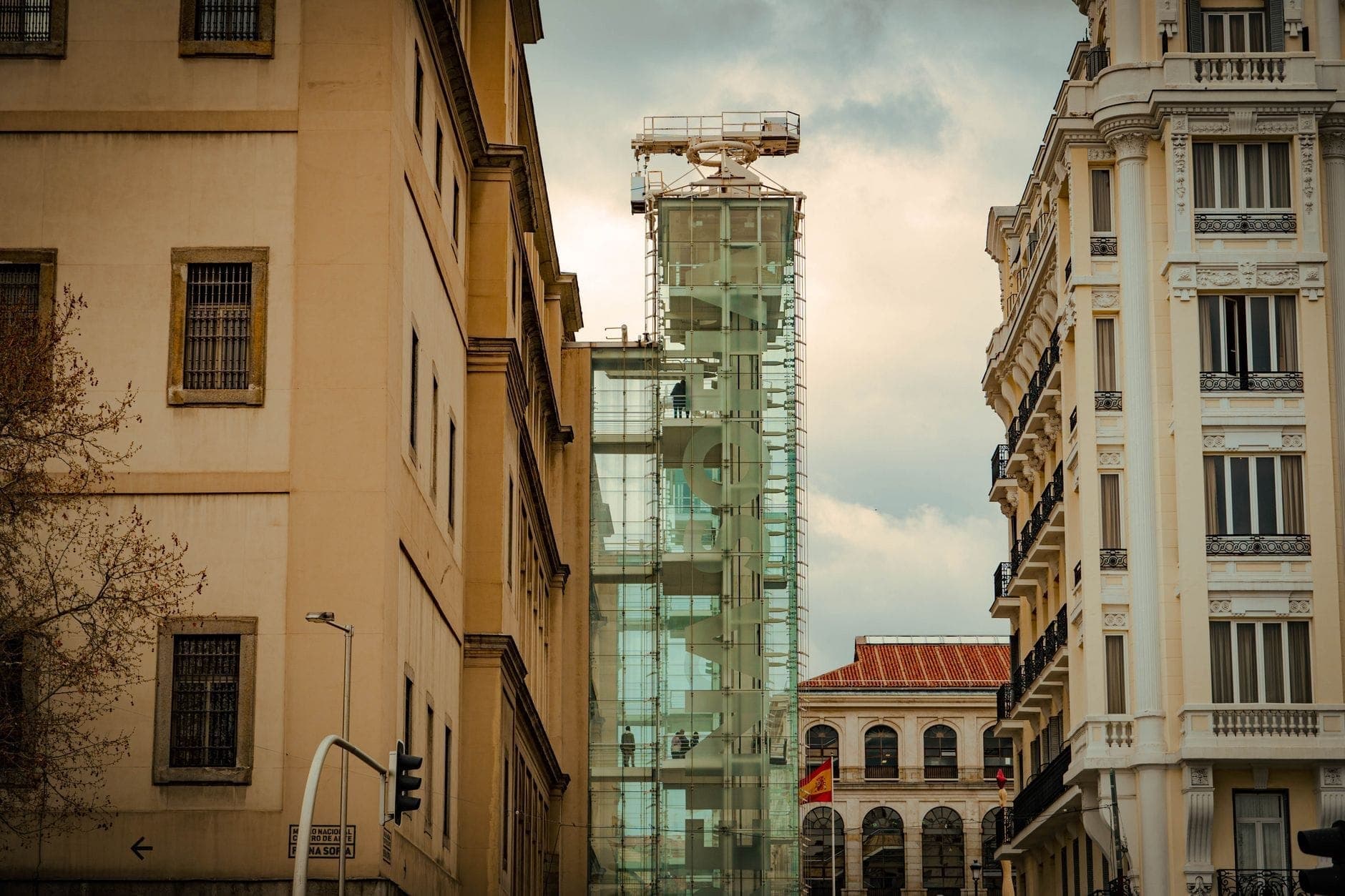 ภาพด้านนอกพิพิธภัณฑ์ Reina Sofía มาดริด โชว์ลิฟต์แก้วไอคอนิคและอาคารประวัติศาสตร์โดยรอบใต้ท้องฟ้าครึ้ม