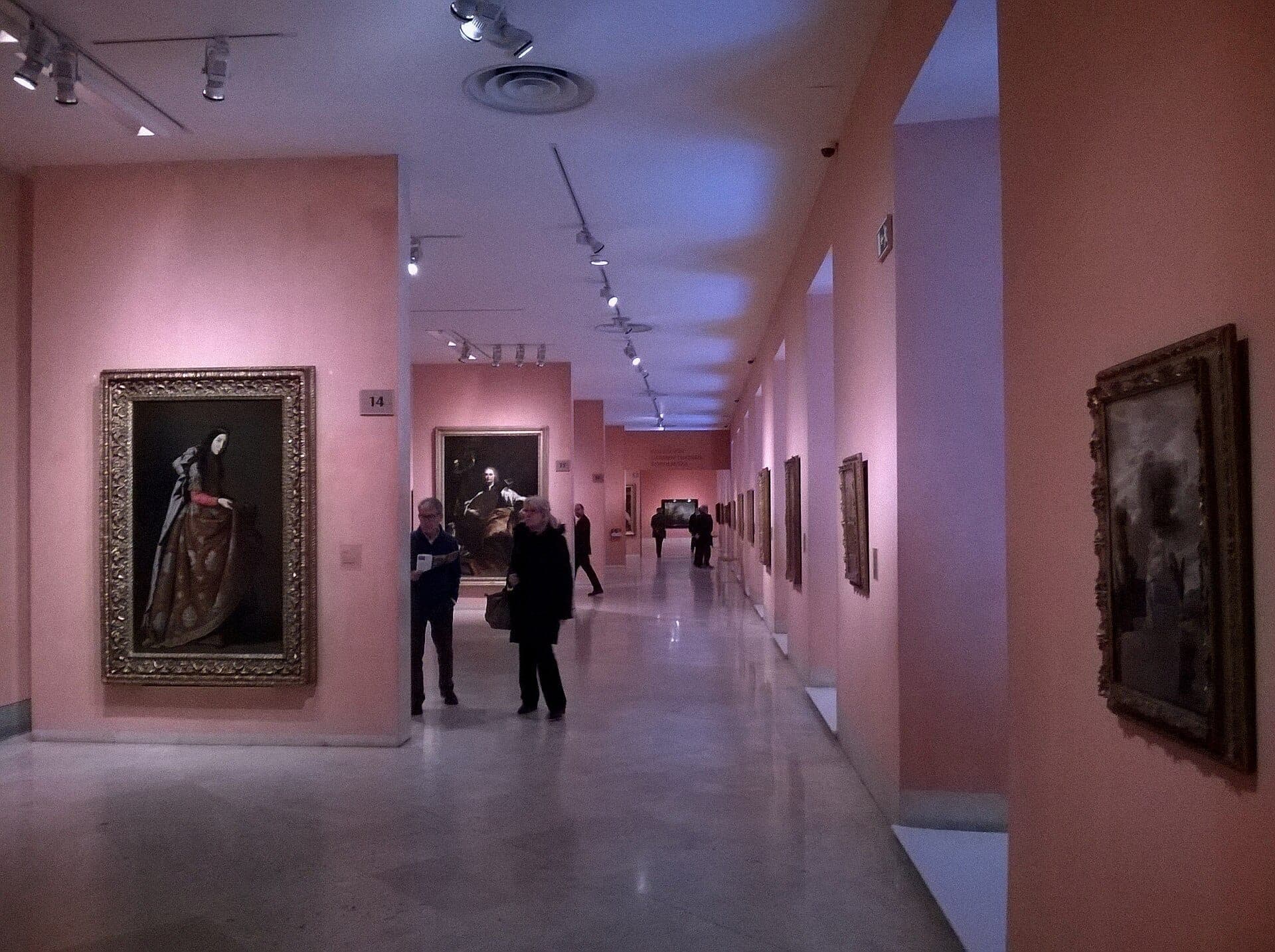 แกลเลอรีภายในของ Museo Thyssen-Bornemisza พร้อมภาพวาดที่มีกรอบ กำแพงสีชมพูอ่อน แสงสปอตไลต์ และผู้เข้าชมที่กำลังชมผลงานศิลปะ