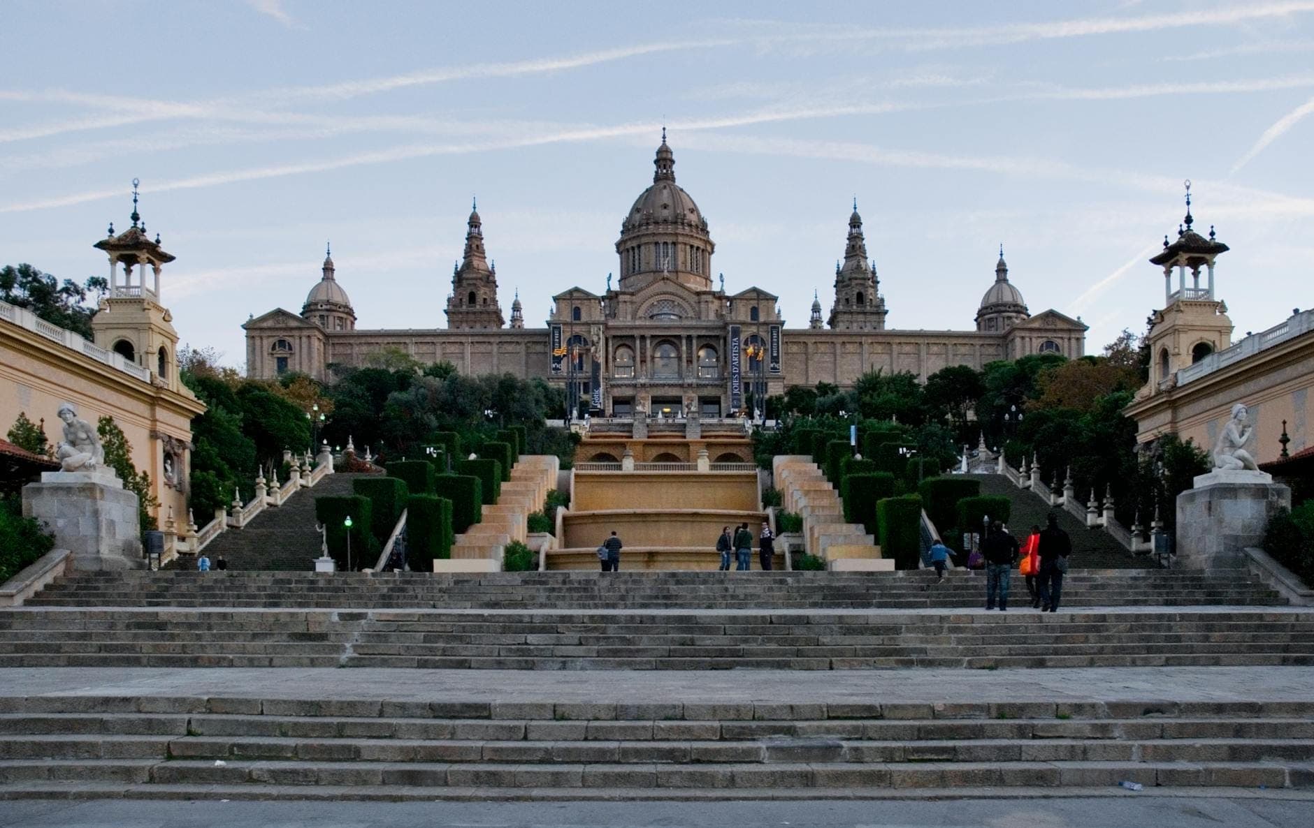 มุมมองกว้างของ Museu Nacional d'Art de Catalunya บนยอดเนินมงต์ฌุยก์ ล้อมรอบด้วยบันไดขนาดใหญ่ สวนสวยงาม และนักท่องเที่ยวที่กำลังเดินขึ้นมา