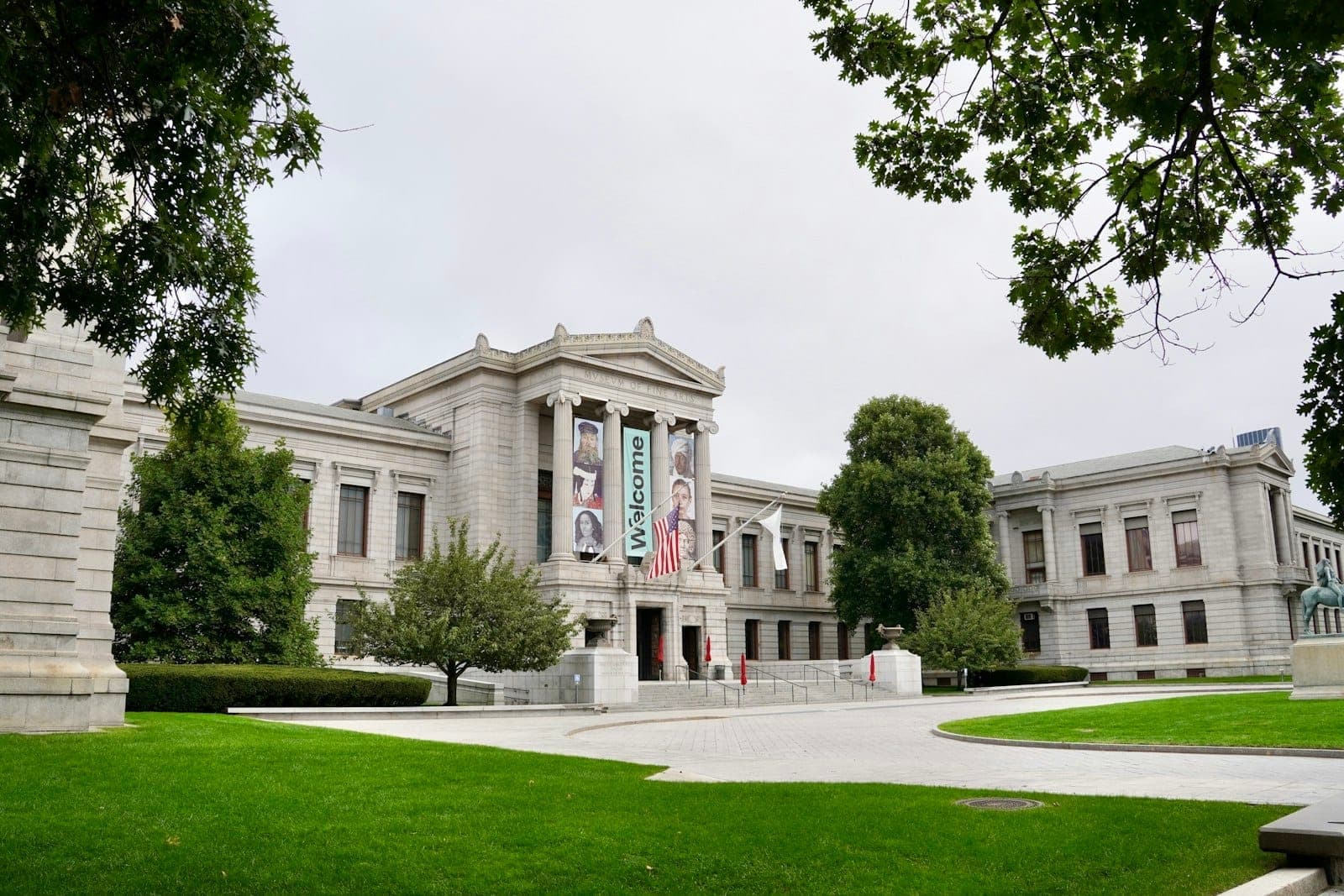 Le Museum of Fine Arts de Boston, avec sa grande façade en pierre, son drapeau américain et ses bannières d'accueil, encadré par de luxuriantes pelouses et des arbres.