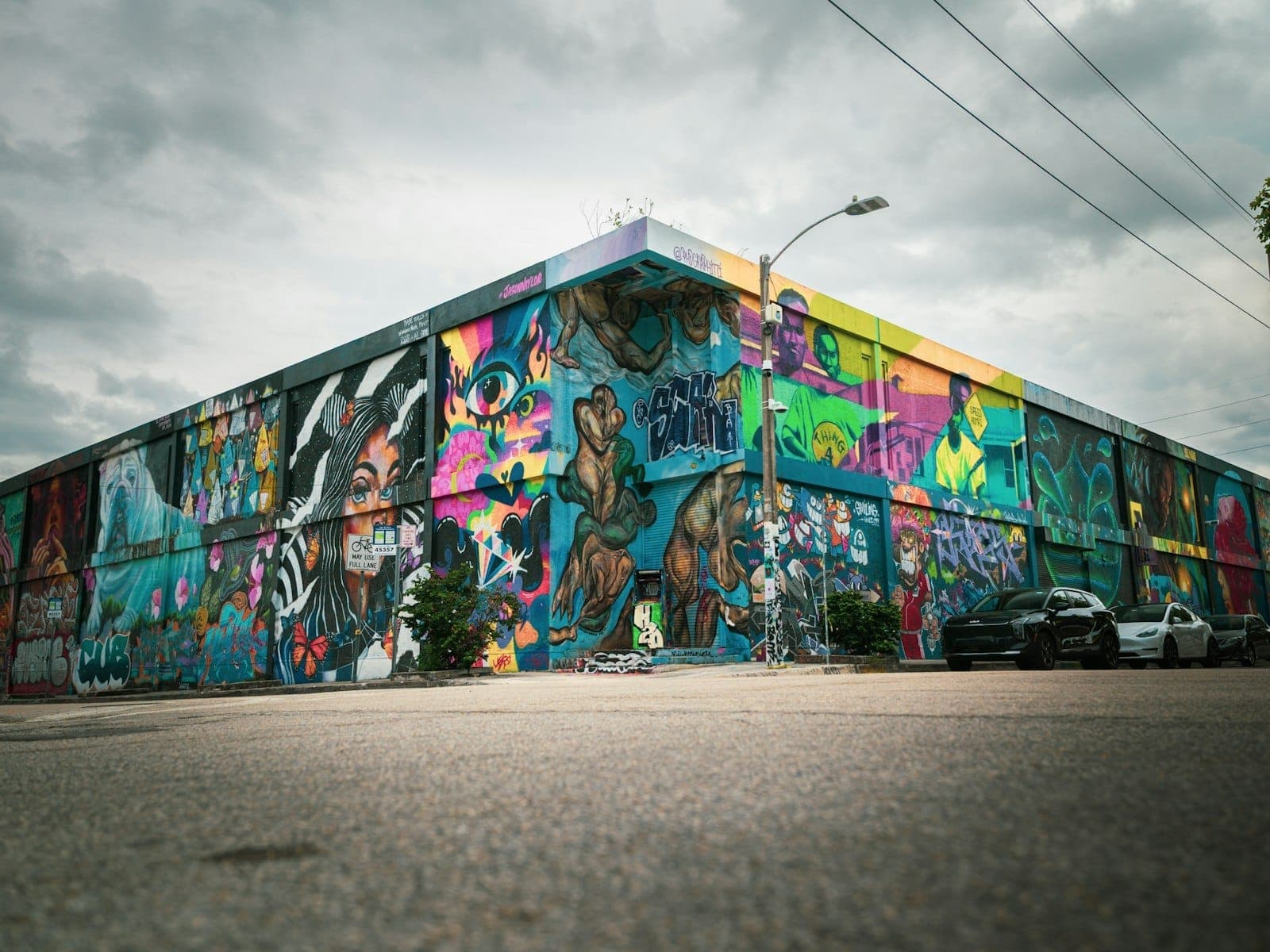 Wide angle view of a building covered with vibrant, large-scale street art murals, set against a dramatic cloudy sky in Miami’s Wynwood district.