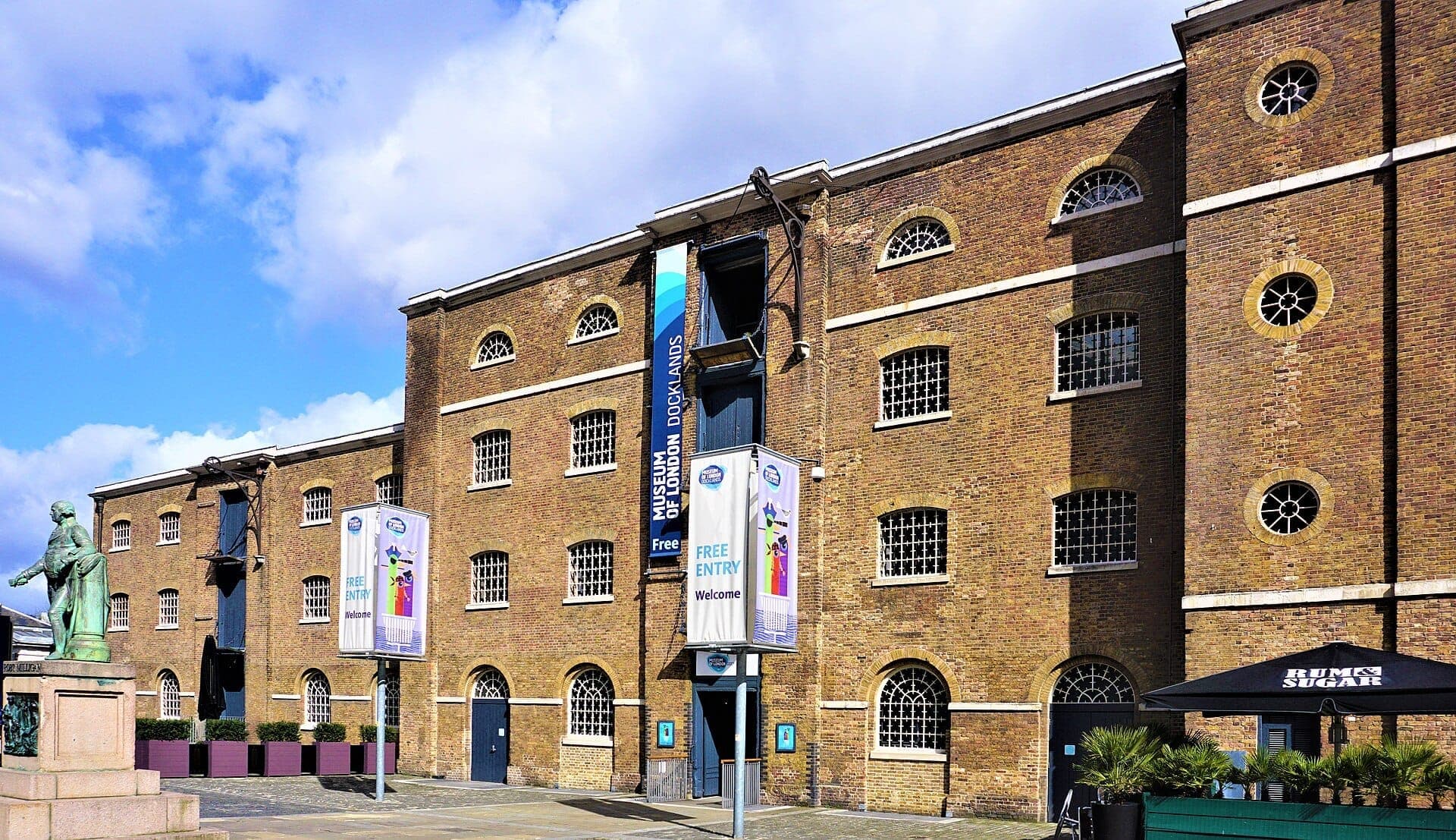 Exterior do London Museum Docklands, um armazém histórico de tijolo com janelas em arco, estátua e faixas para visitantes sob um céu azul brilhante.