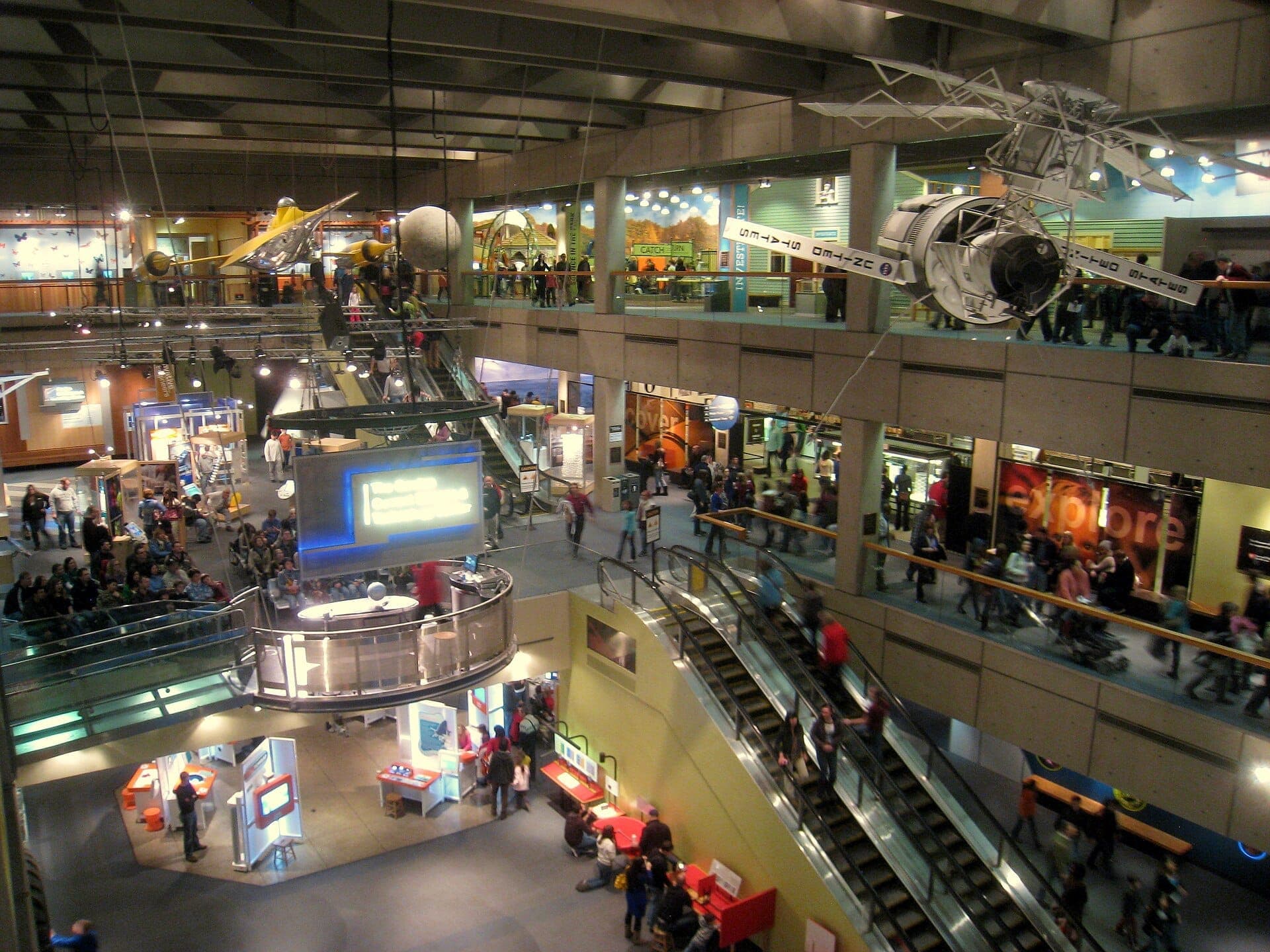 Vue panoramique de l'intérieur du Museum of Science de Boston, montrant des expositions interactives, des escalators et des maquettes de vaisseaux spatiaux suspendues, avec des visiteurs explorant plusieurs niveaux.