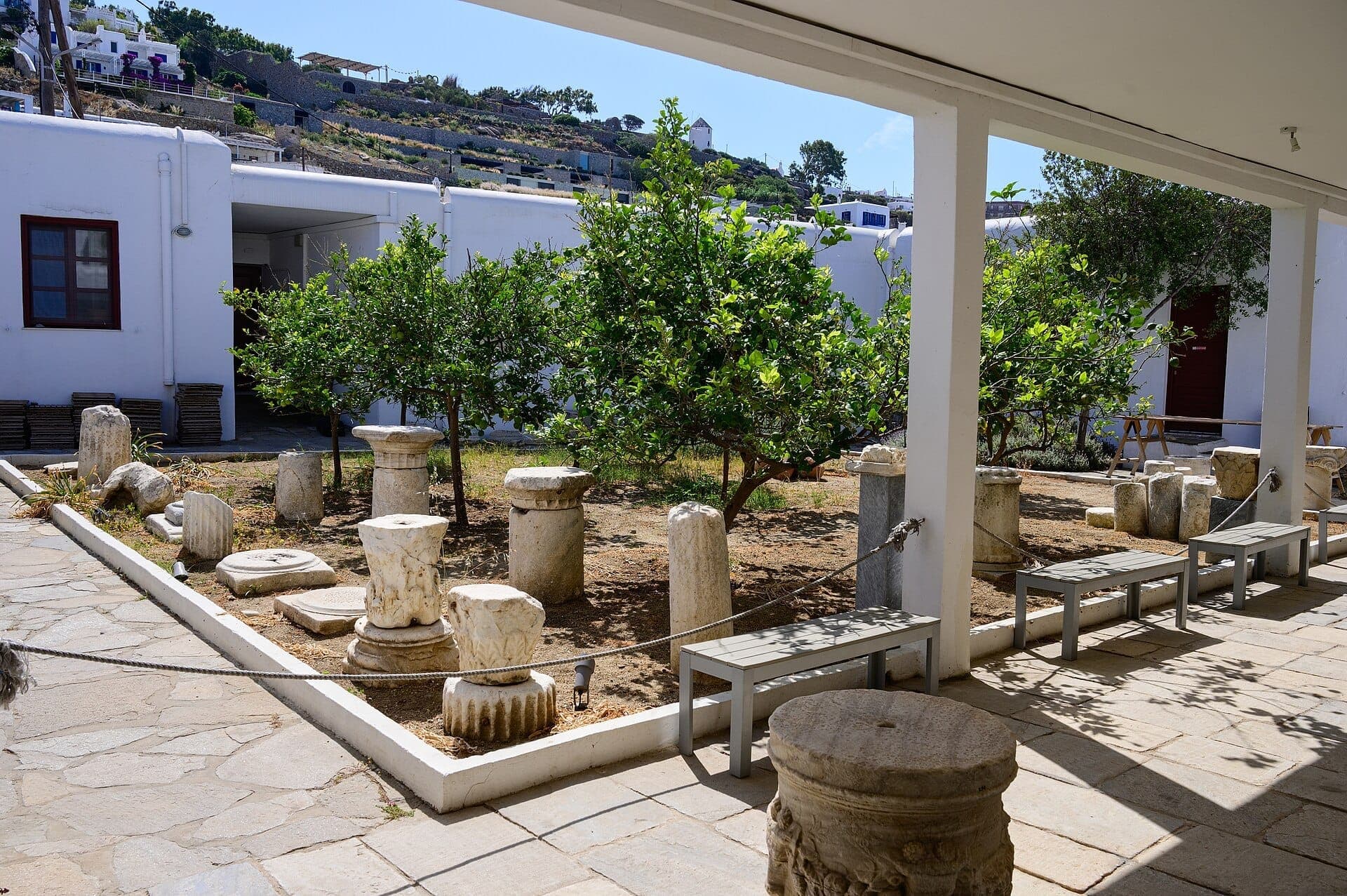 Antike Marmorsäulen und Steinartefakte, ausgestellt im sonnigen Innenhof des Archäologischen Museums von Mykonos, umgeben von weiß getünchten Gebäuden und Grünpflanzen.
