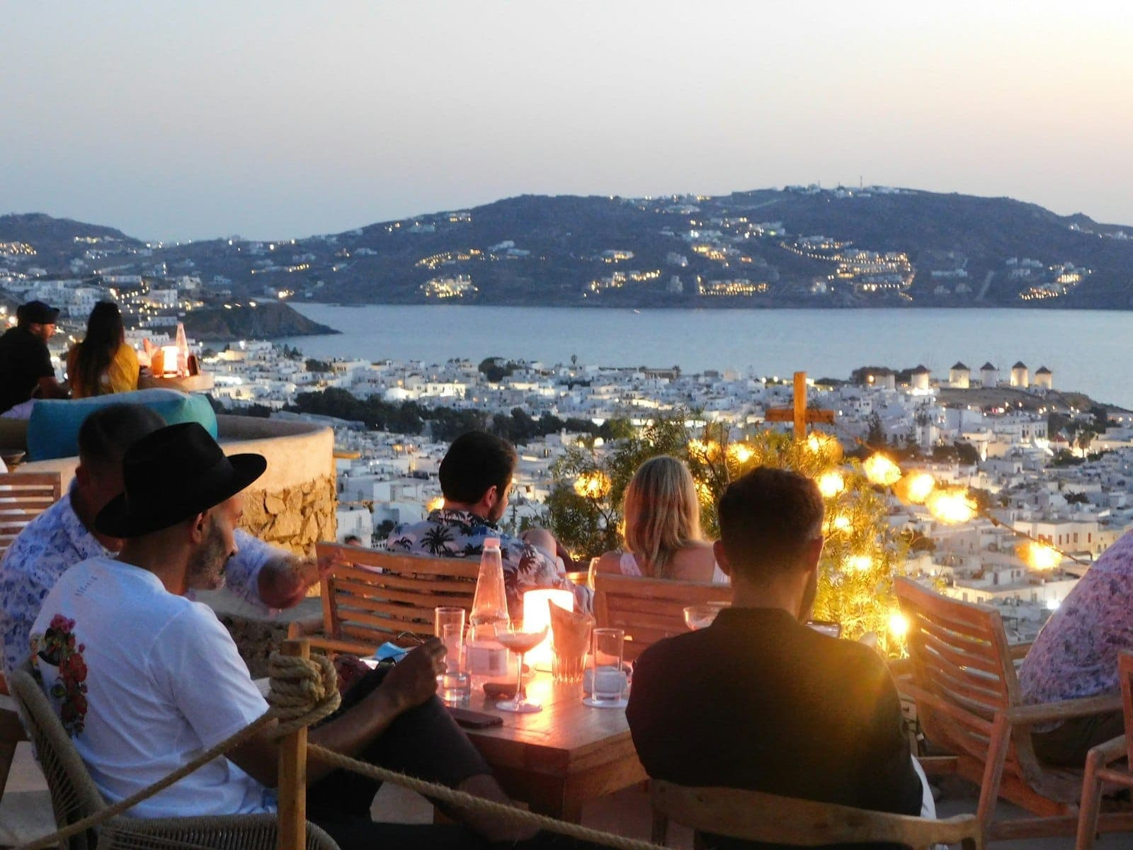 Menschen genießen Sundowner-Drinks auf einer Außenterrasse mit Blick auf Mykonos Town, die Ägäis und die berühmten Windmühlen.