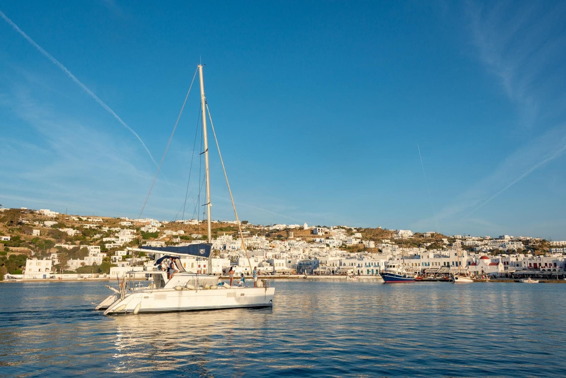 Segelboot in den Gewässern vor Mykonos mit weiß getünchten Stadtgebäuden und Hügeln im Hintergrund unter einem klaren blauen Himmel.