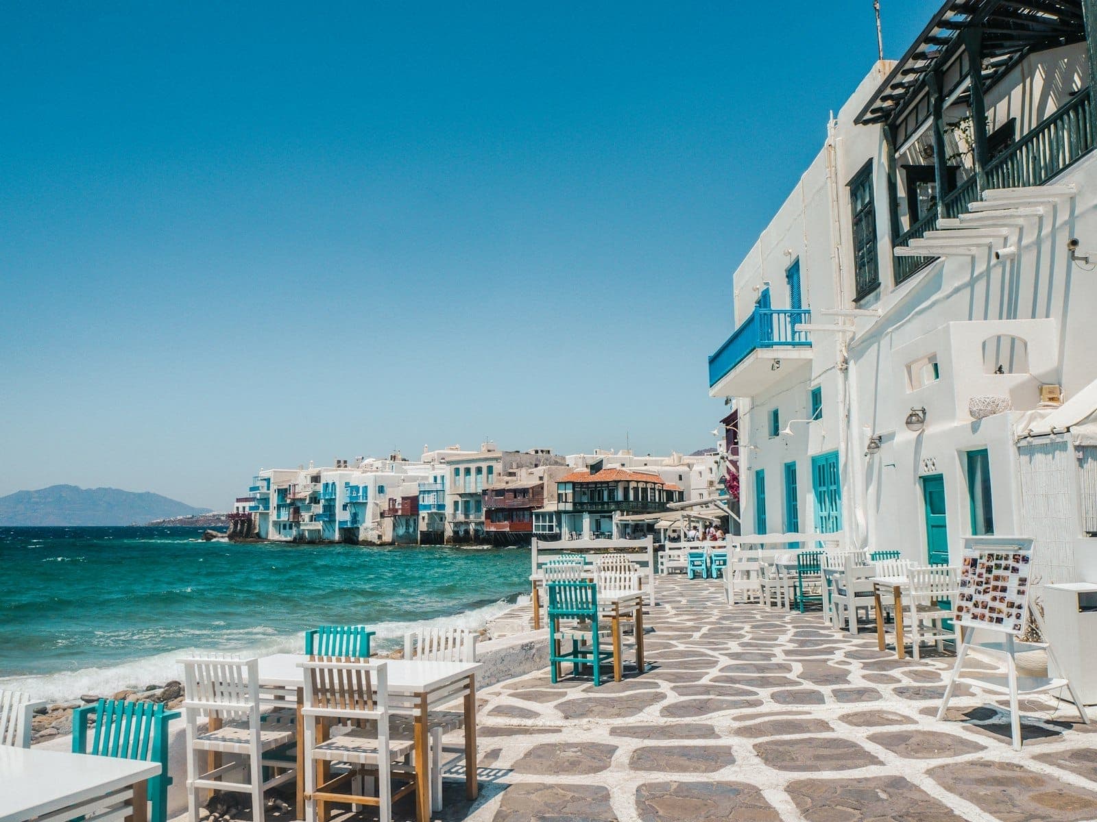 Meerblick in Mykonos mit weiß getünchten Gebäuden, Tischen und Stühlen am Wasser und dem ikonischen Viertel Little Venice im Hintergrund.