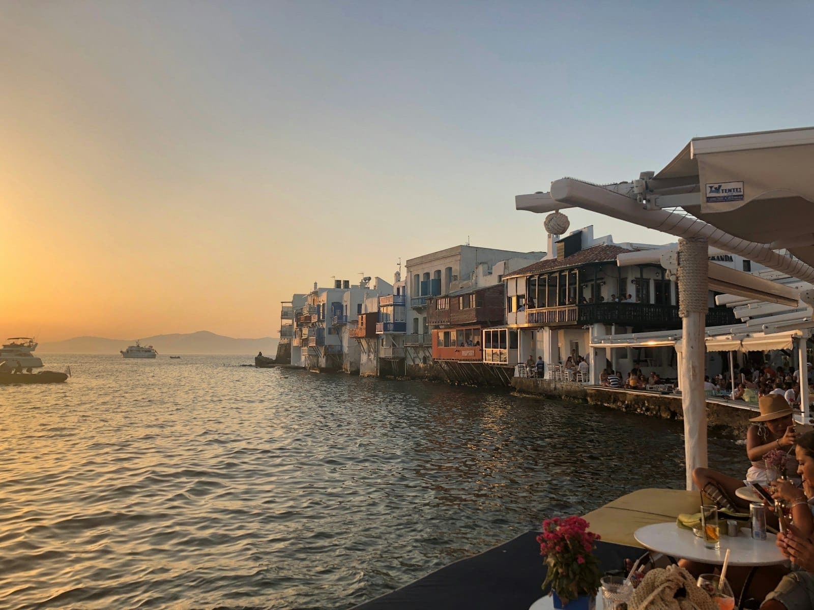 Restaurants am Meer und bunte Häuser von Mykonos' Kleinem Venedig bei Sonnenuntergang, mit Tischen mit Blick auf das Wasser und einem romantischen orangefarbenen Himmel.