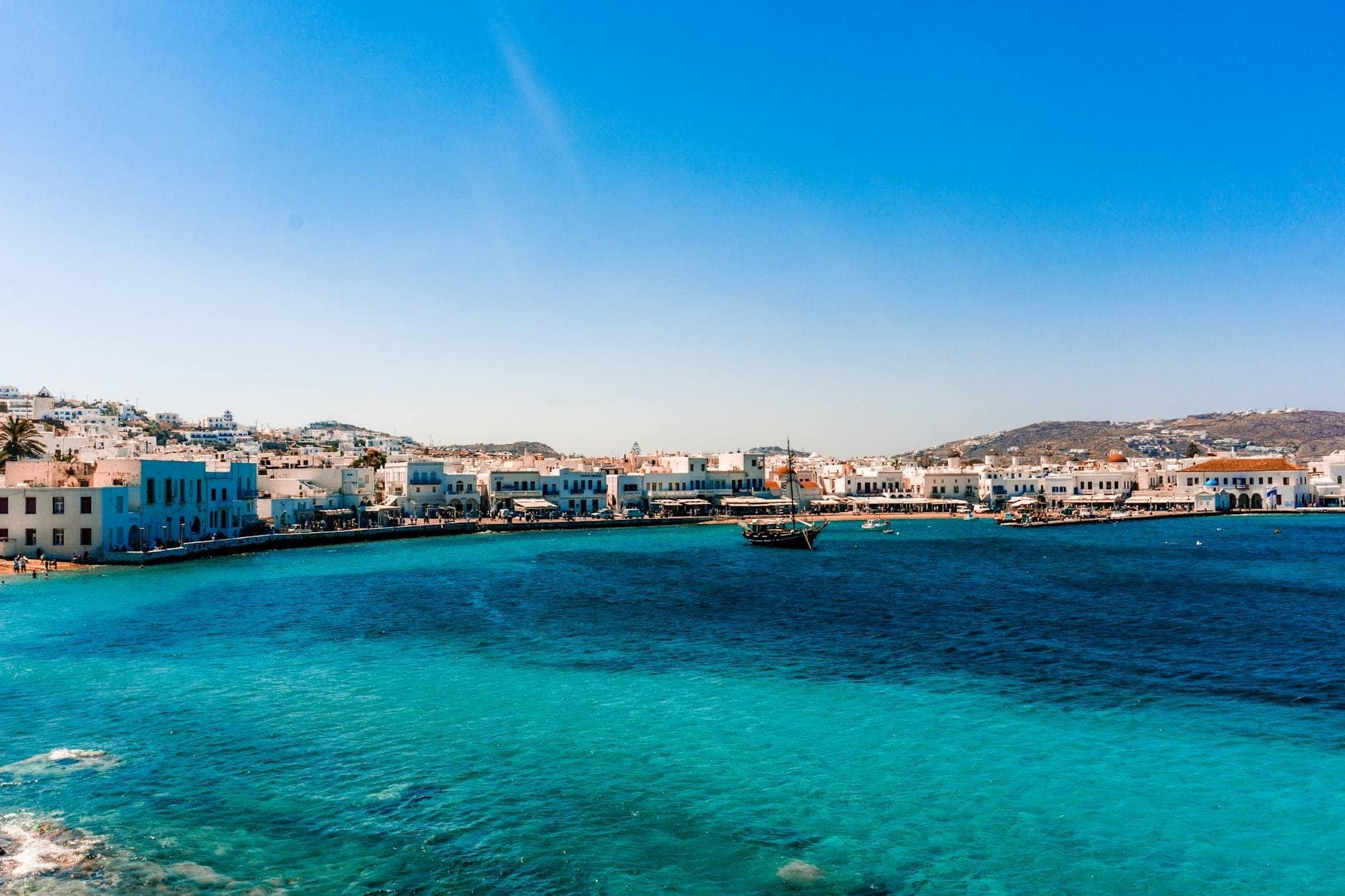 Strahlend blaues Ägäisches Meer mit Luxusbooten und dem Uferpromenade der Stadt Mykonos, weiß getünchten Gebäuden und Hügeln im Hintergrund unter einem klaren Himmel.