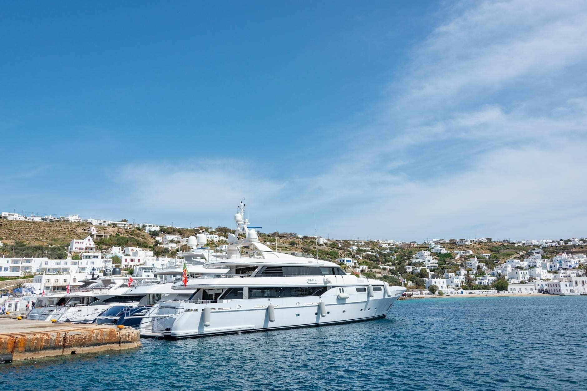 Große Luxusyacht in einem Hafen von Mykonos, mit kristallklarem blauem Wasser, weißgetünchten Gebäuden und sanften Hügeln unter strahlend blauem Himmel.
