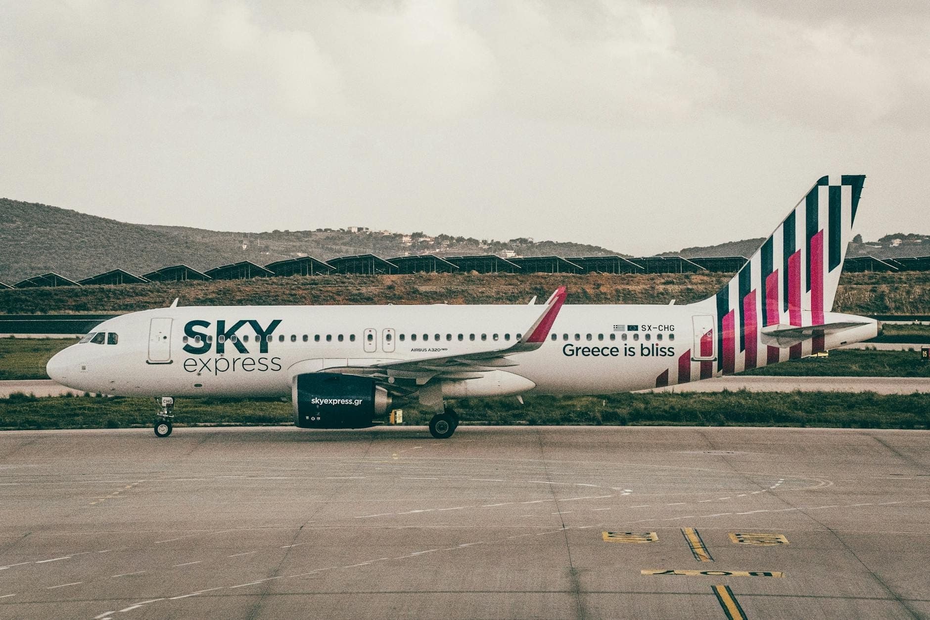 Sky Express-Flugzeug mit dem Slogan 'Greece is bliss' auf dem Rollfeld eines griechischen Inselflughafens, umgeben von Hügeln und bewölktem Himmel.