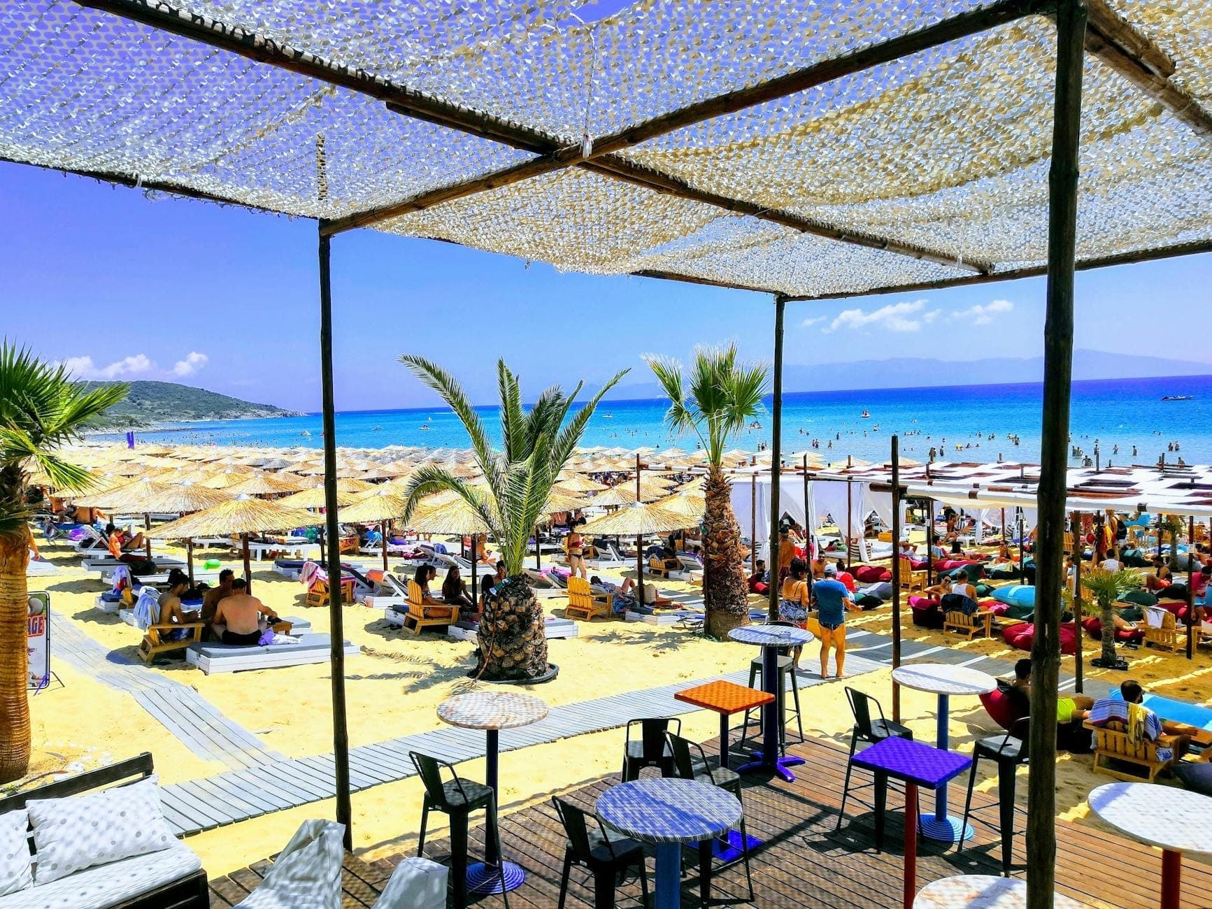 Eine lebhafte Beach-Club-Szene in Mykonos mit Sonnenliegen, Sonnenschirmen, entspannenden Menschen und einem klaren Blick auf das türkisfarbene Meer.