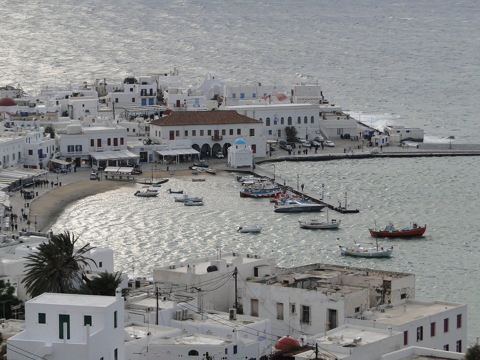 Panoramablick auf den Alten Hafen von Mykonos mit weiß getünchten Gebäuden, kleinen im Hafen vertäuten Booten und der sanften Kurve des Strandbereichs am Wasser.