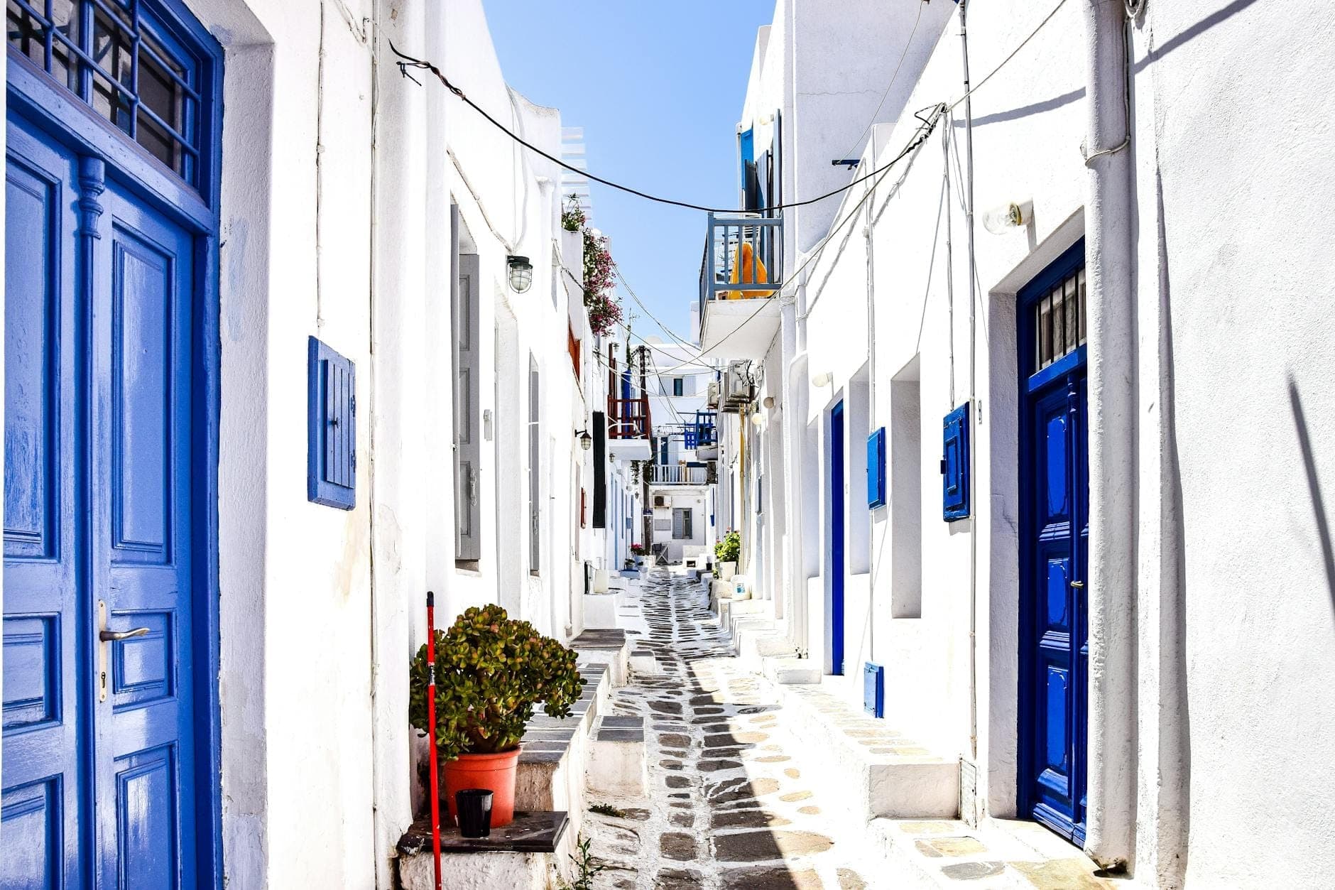 Helle, sonnenbeschienene Steingasse in der Altstadt von Mykonos mit traditionellen weißen Gebäuden und blauen Türen, einladend für einen malerischen Spaziergang.